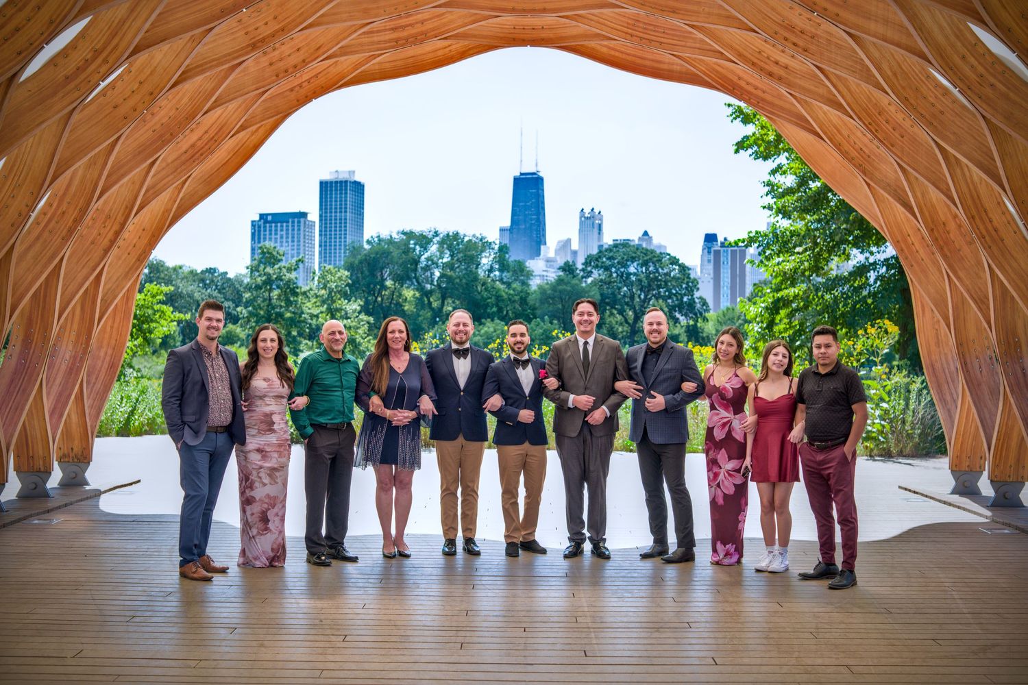 Wedding party lineup under a curved wooden structure with city skyline in background.