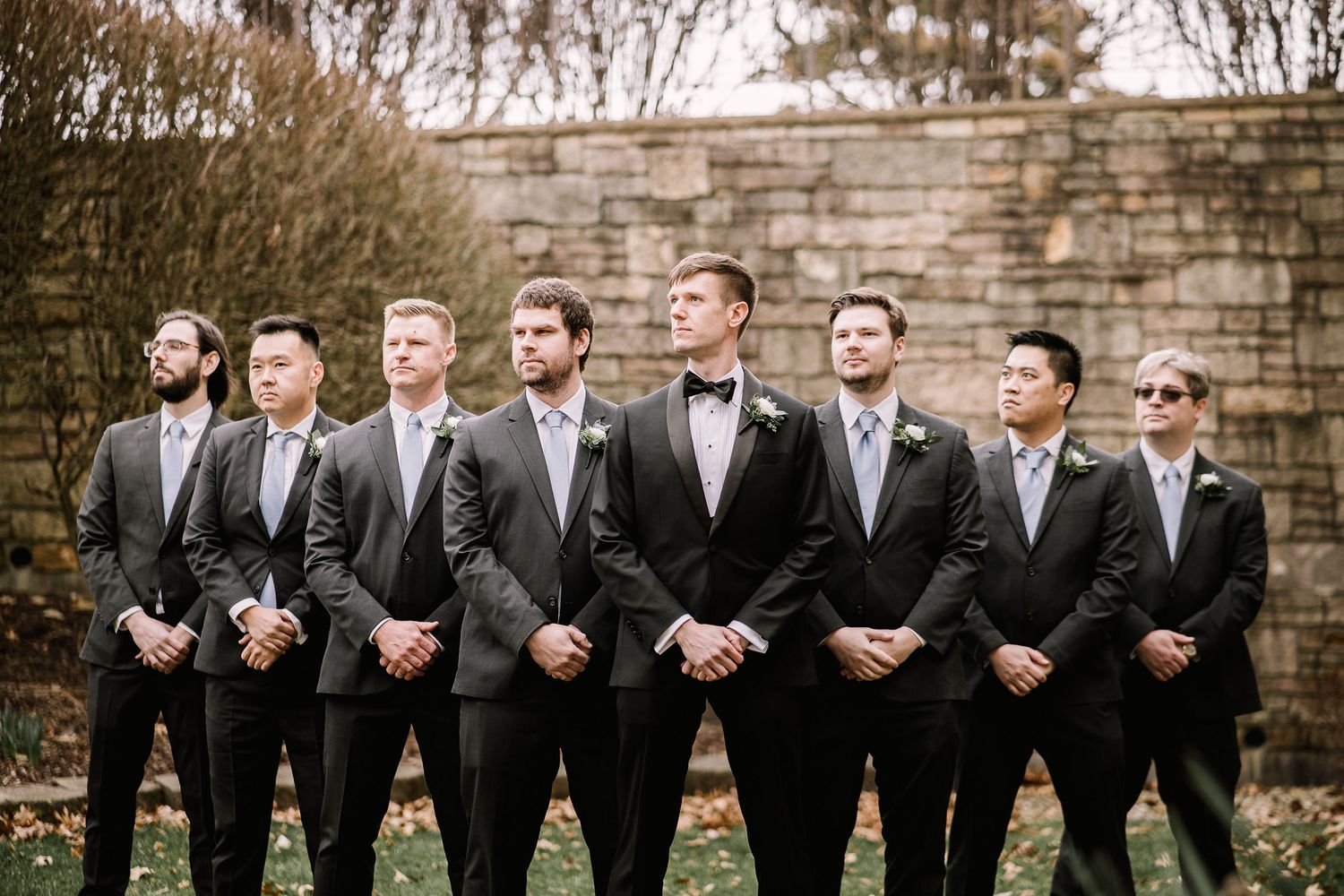 Wedding party in formal black suits lined up against stone wall for group photo.
