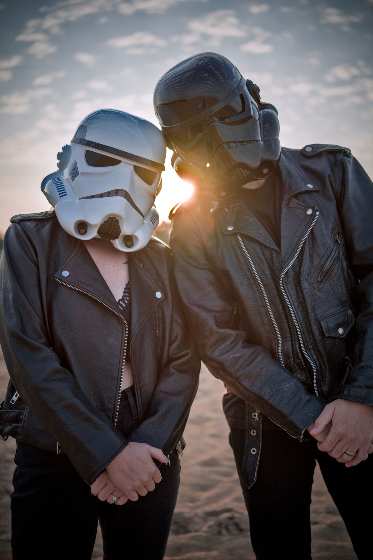 Two people wearing Star Wars stormtrooper helmets and black leather jackets pose at sunset on a sandy beach.