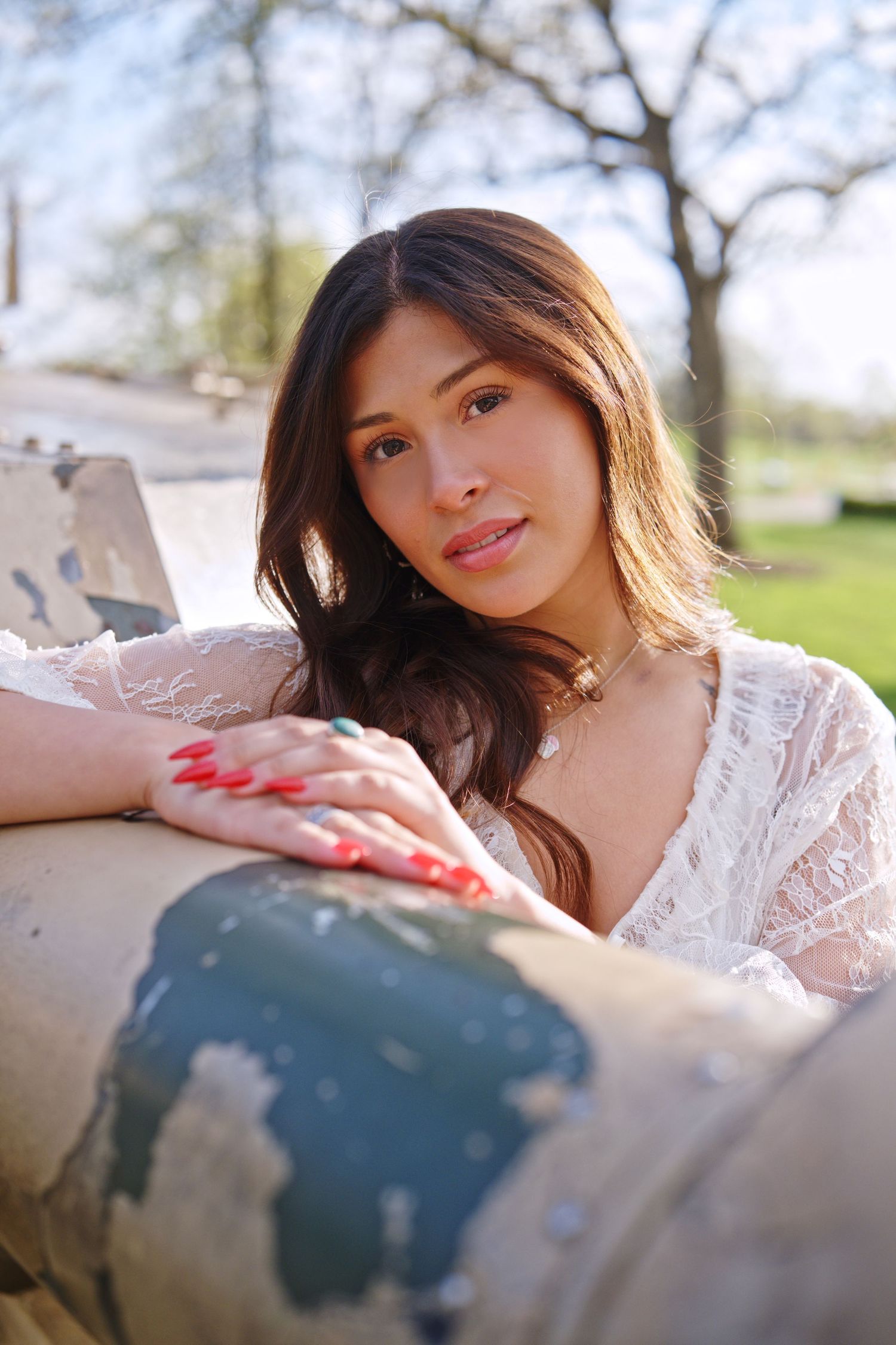 Young woman with red nails and lace top resting on weathered surface outdoors in sunny park setting.