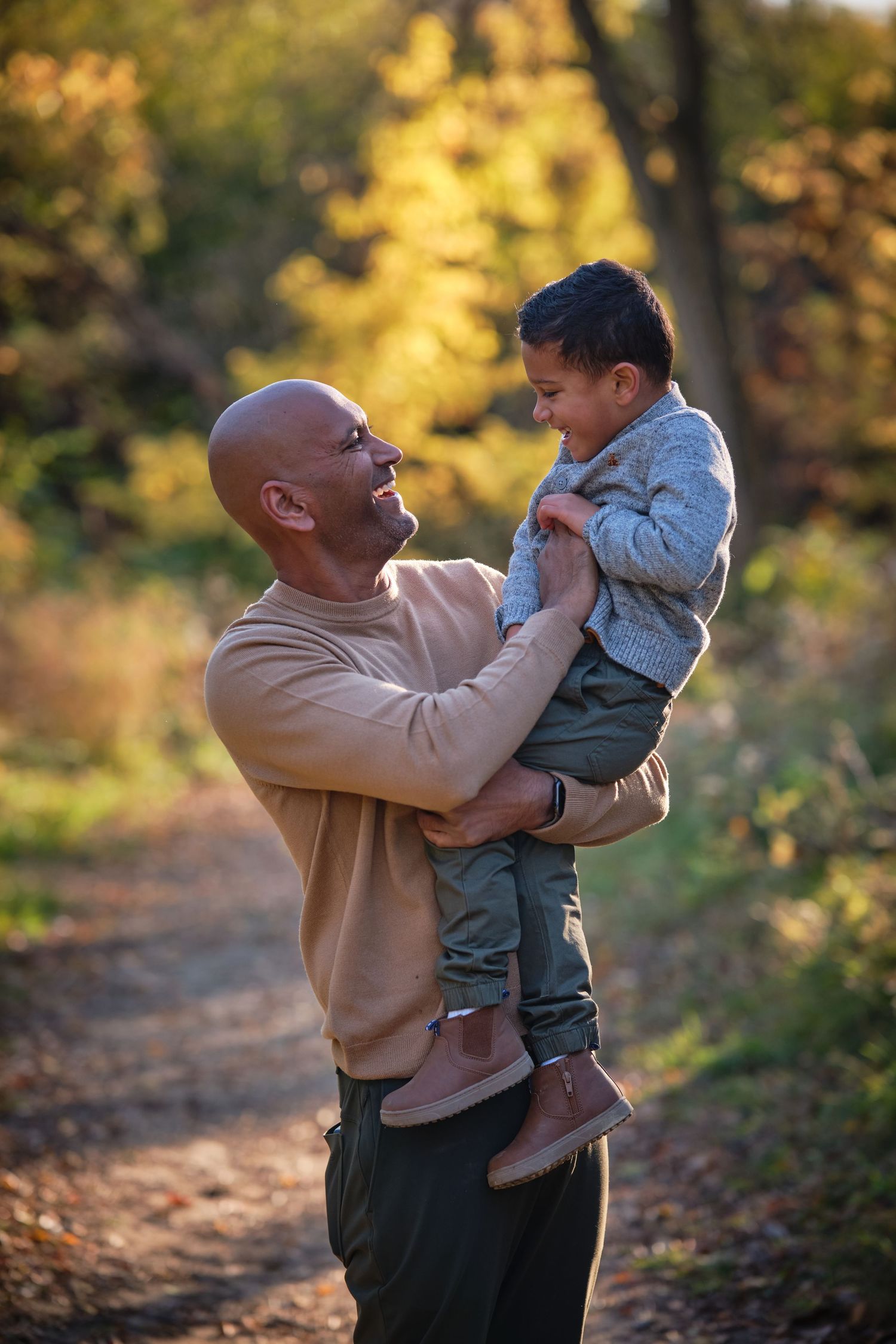 Bald man in beige sweater joyfully lifts young child on a sunlit autumn forest path.