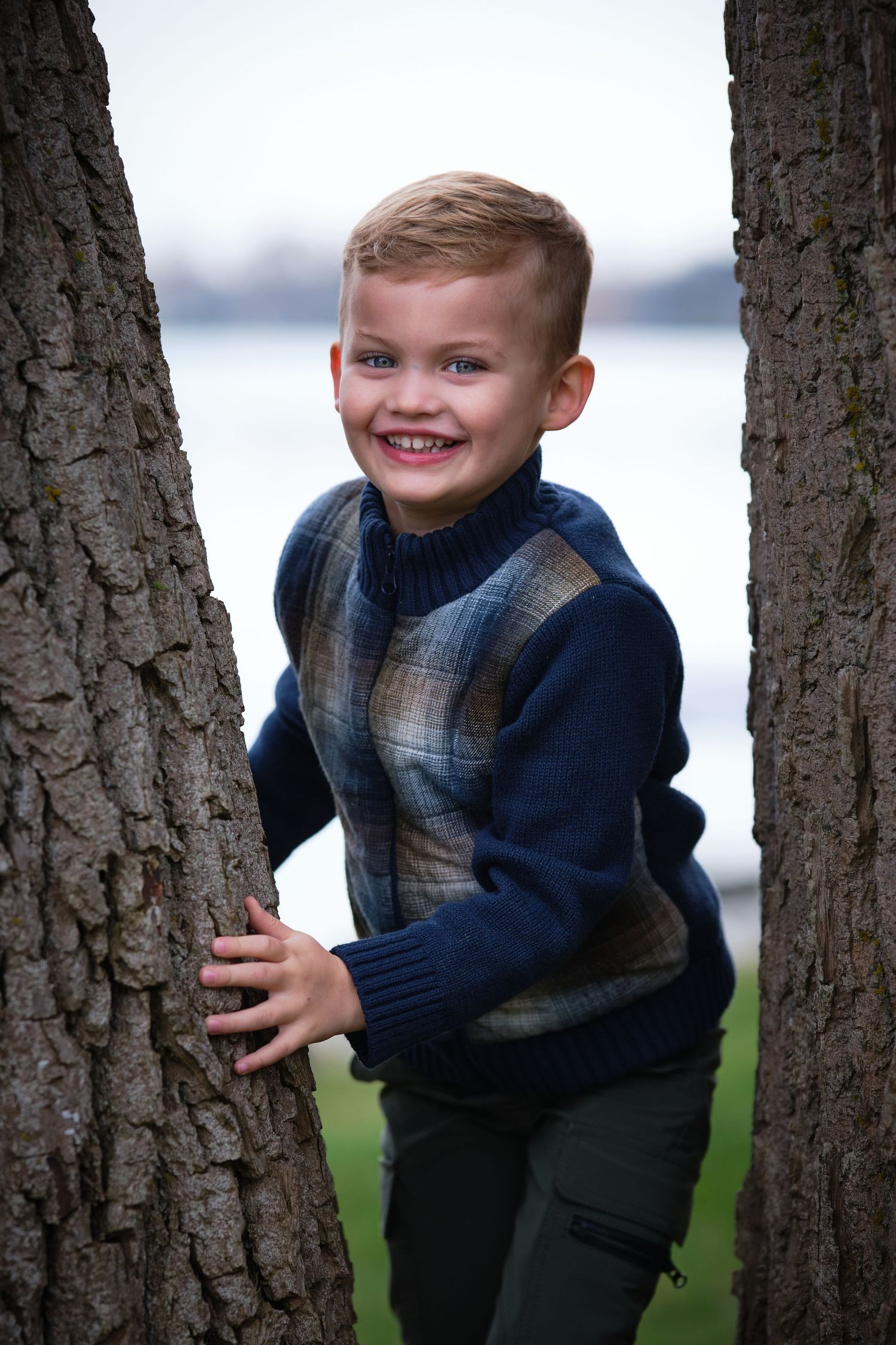 Smiling young boy in a plaid sweater standing between two tree trunks near a waterfront.