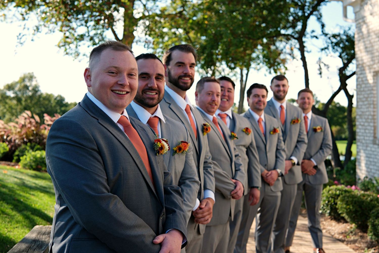 Groomsmen in gray suits and coral ties pose outdoors for wedding photos.