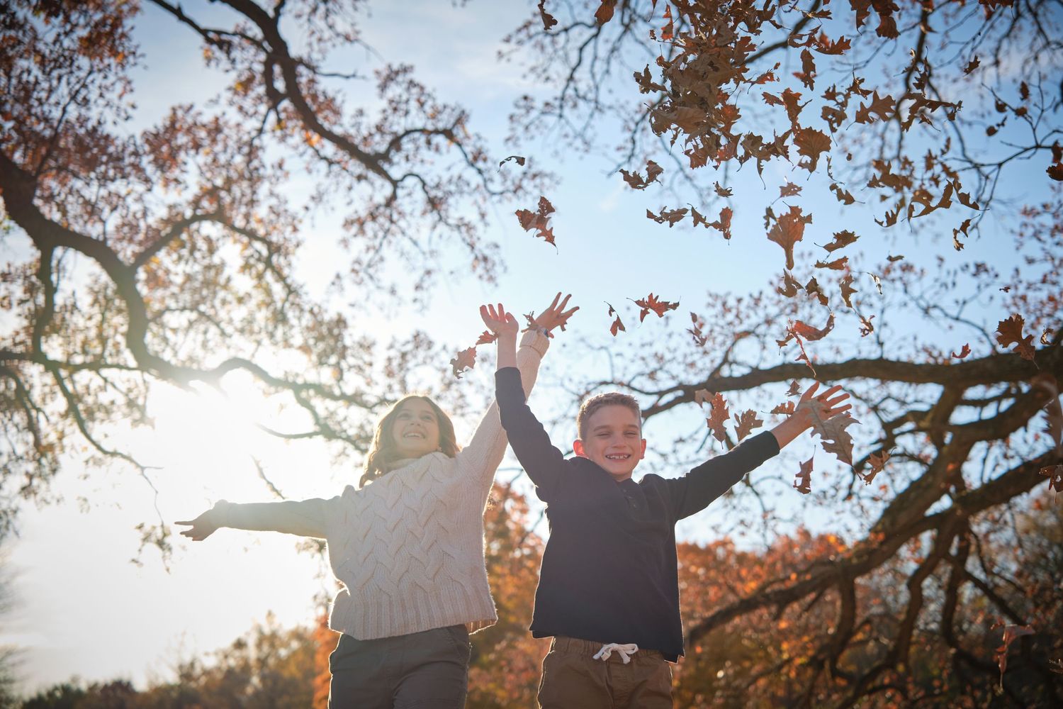 Two children joyfully tossing autumn leaves in the air under a sunlit tree with colorful fall foliage.