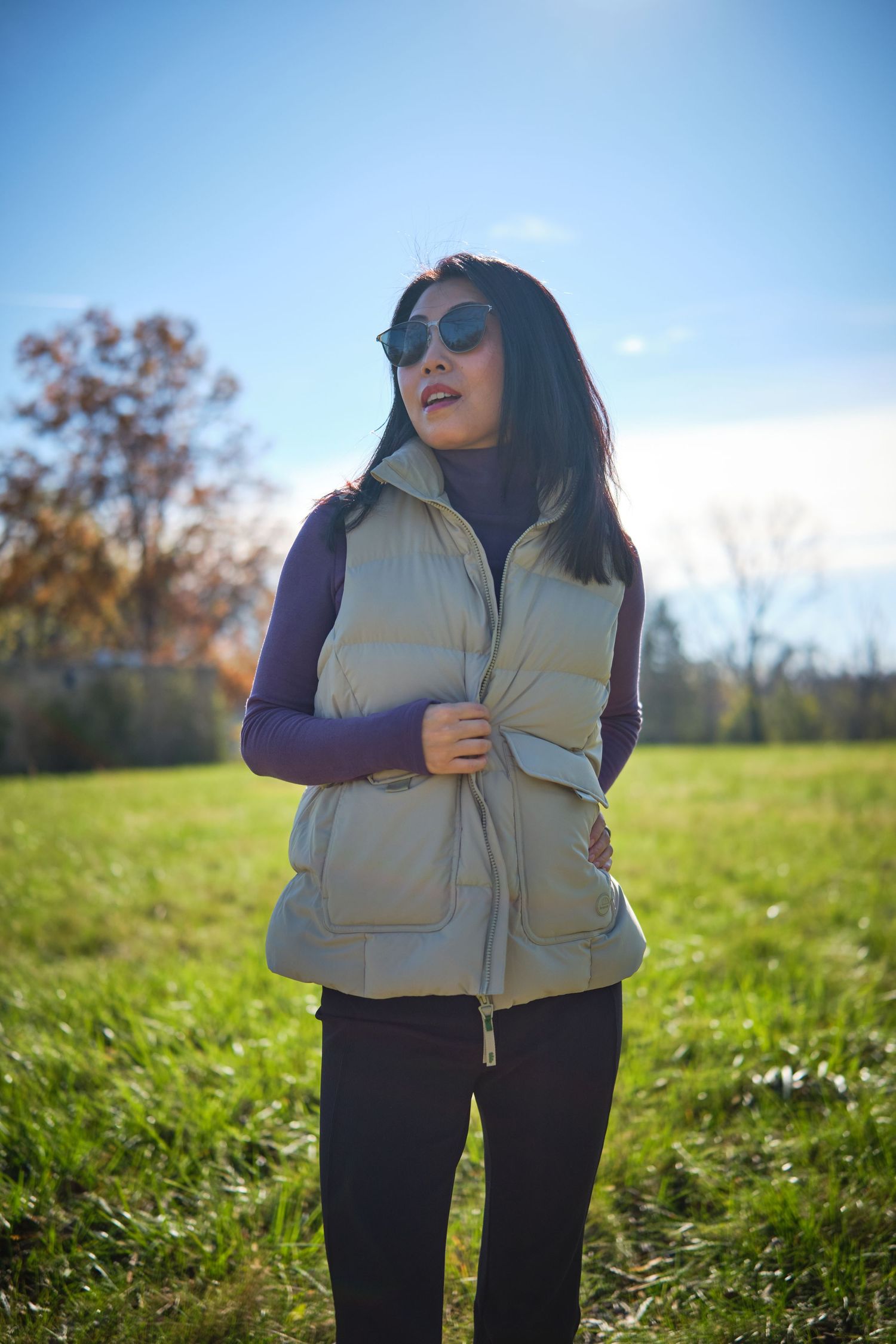 Woman in beige puffer vest and purple turtleneck wearing sunglasses standing in green field on sunny day.