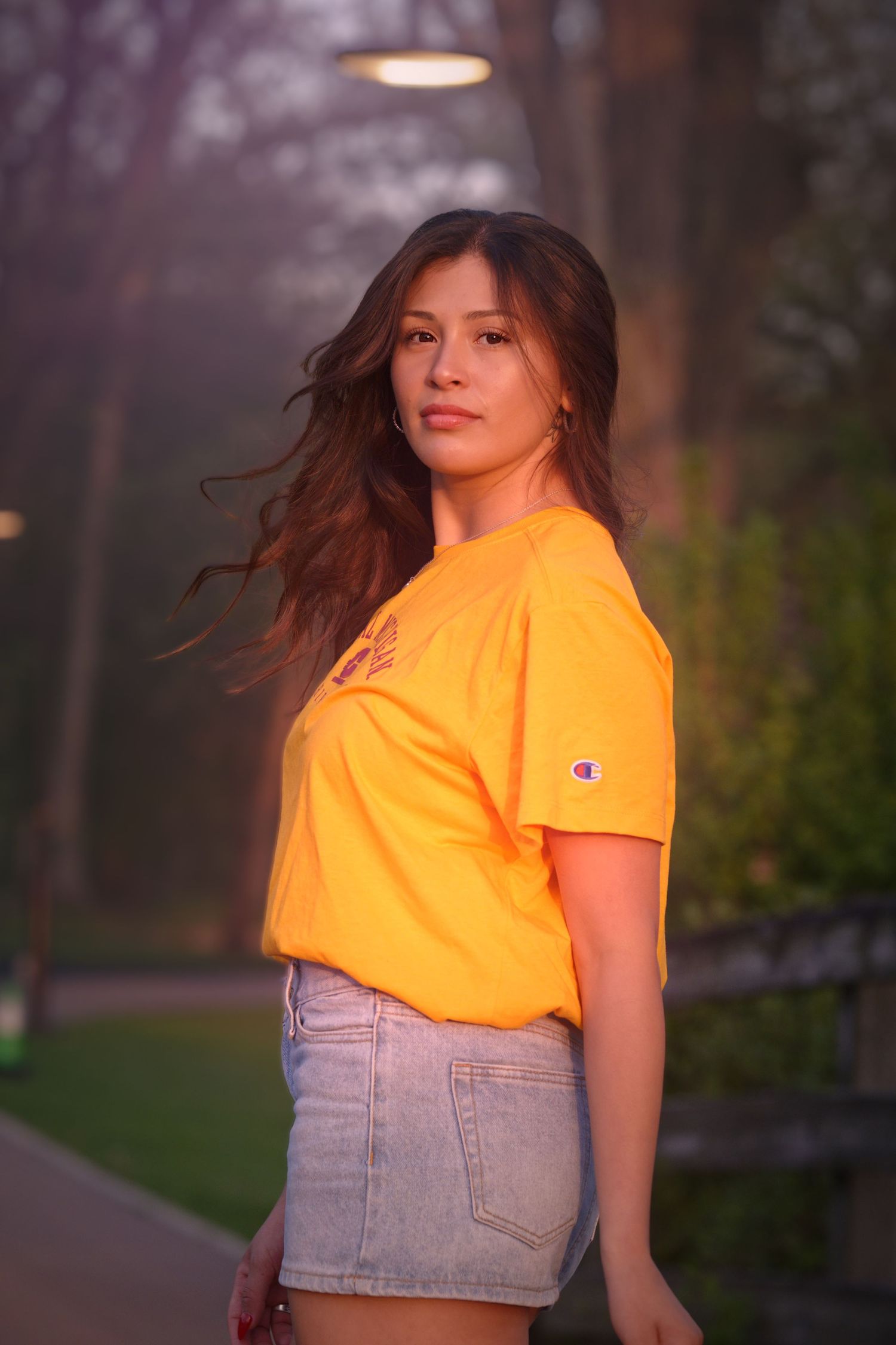 Young woman in orange Champion t-shirt and denim skirt posing outdoors in a park at golden hour.