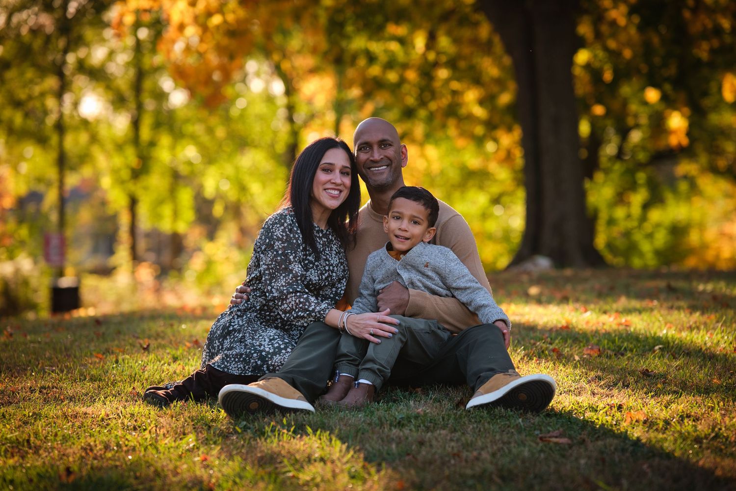 Family of three sitting on grass in autumn park, smiling, with golden foliage trees in background.