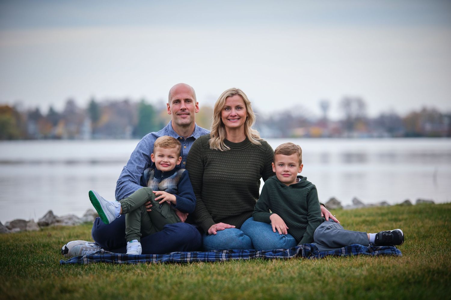 Family of four smiling together on a plaid blanket by a lake on a cloudy autumn day.