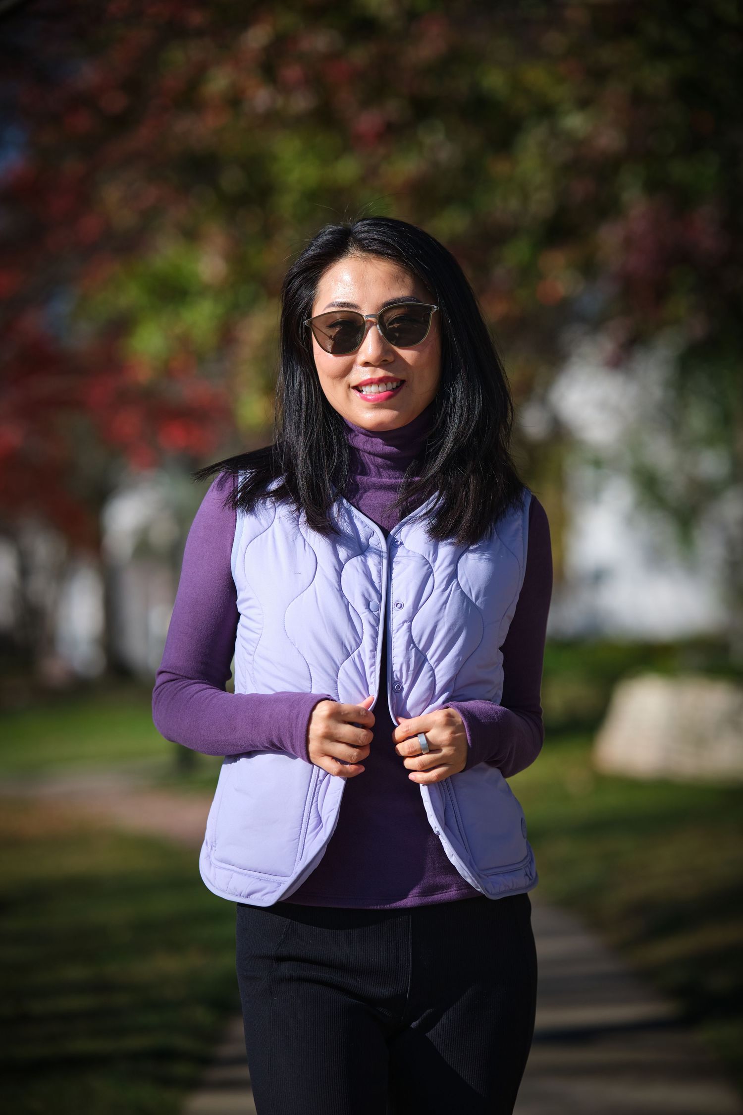 Woman in purple turtleneck and lavender puffer vest wearing sunglasses, smiling outdoors in autumn park.