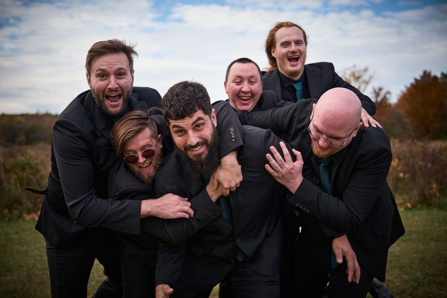 Groomsmen share a candid laughing moment together outdoors in black suits during a wedding photoshoot.