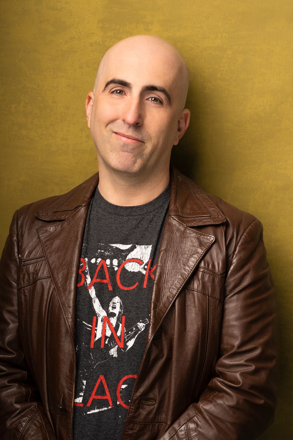 Smiling person in brown leather jacket and rock band t-shirt against gold background.