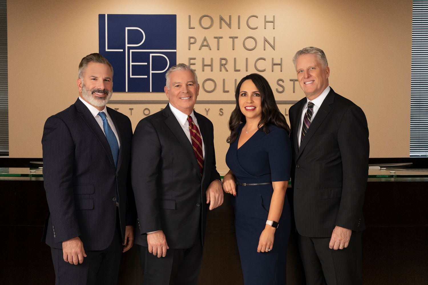 Four business professionals in formal attire stand smiling together in front of a law office logo and wall.