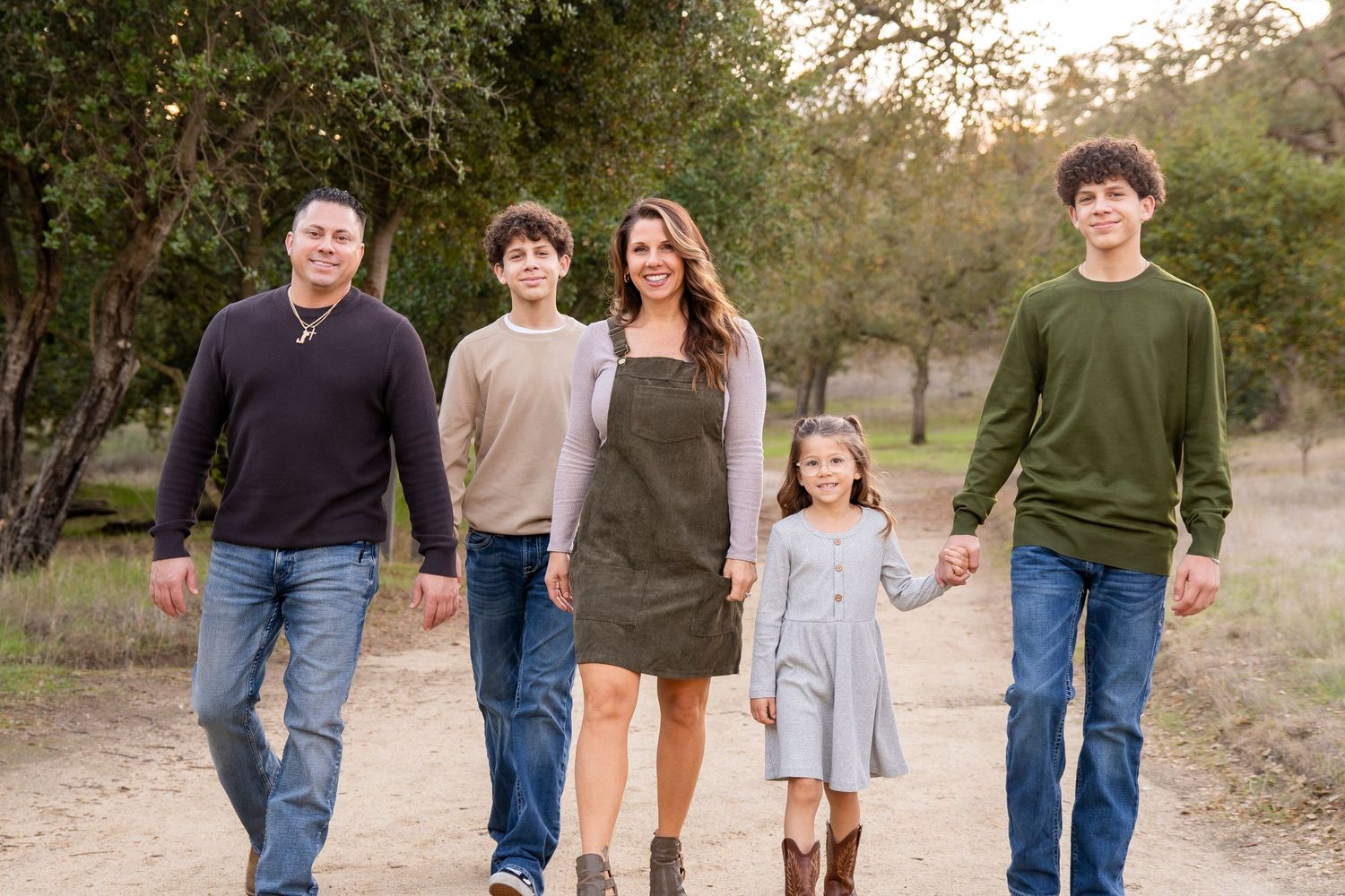 Family of five walking outdoors on a dirt path surrounded by trees, smiling and holding hands.