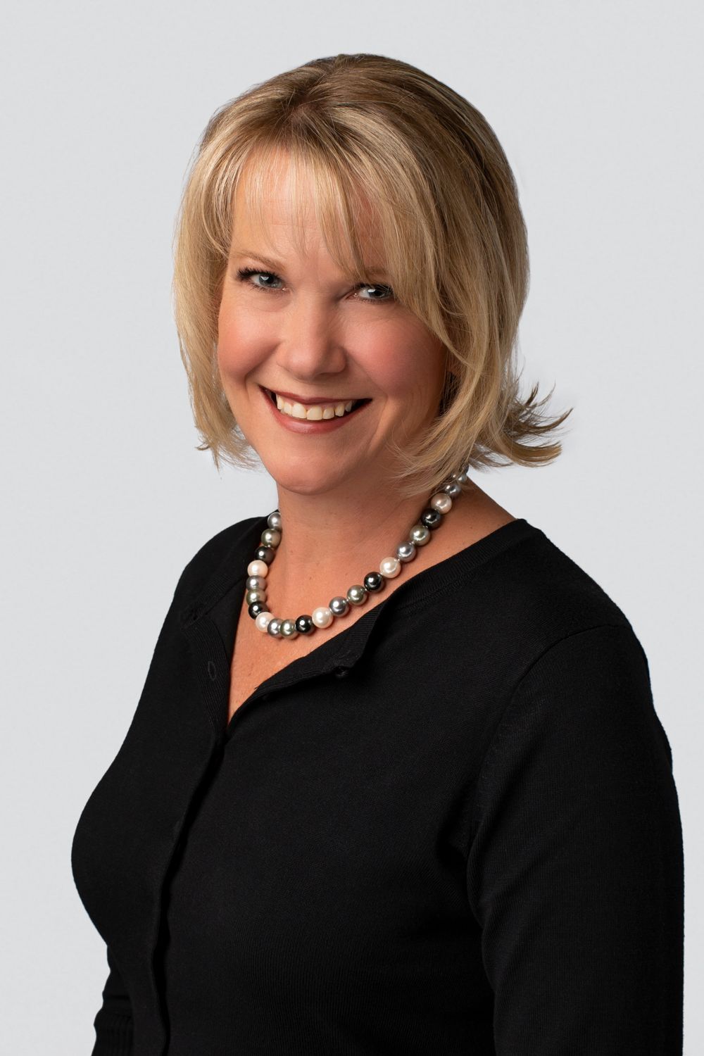 Professional headshot portrait of a woman in black top with a statement necklace against white background.
