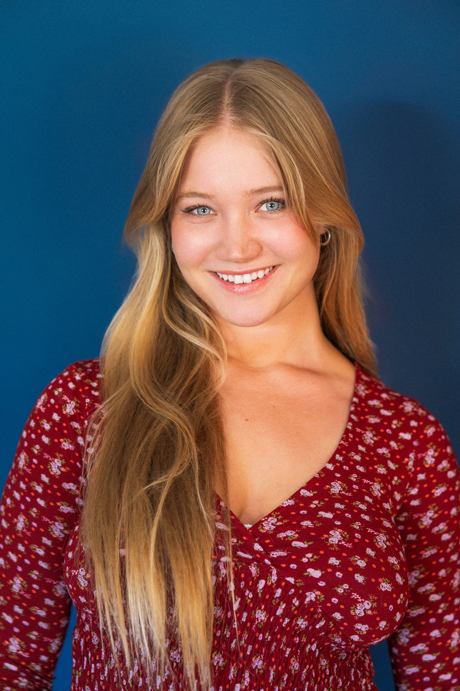 Young woman in red floral v-neck top smiles warmly against blue background.
