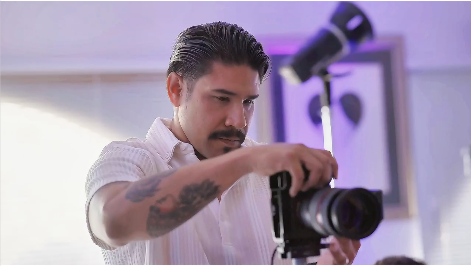 Tattooed male photographer adjusting camera on tripod in bright studio setting.