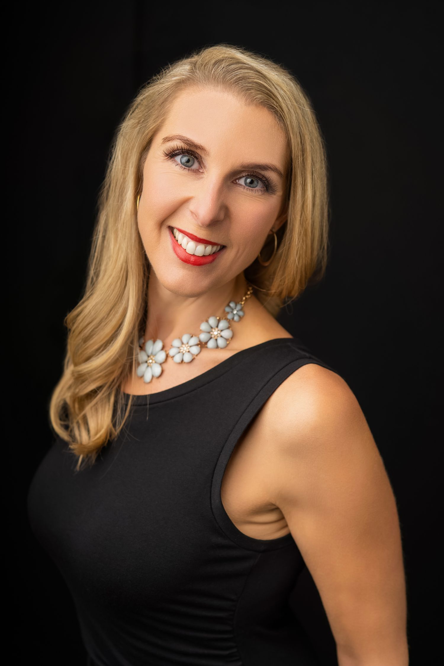 Studio portrait of woman in black sleeveless top with floral necklace against dark background.