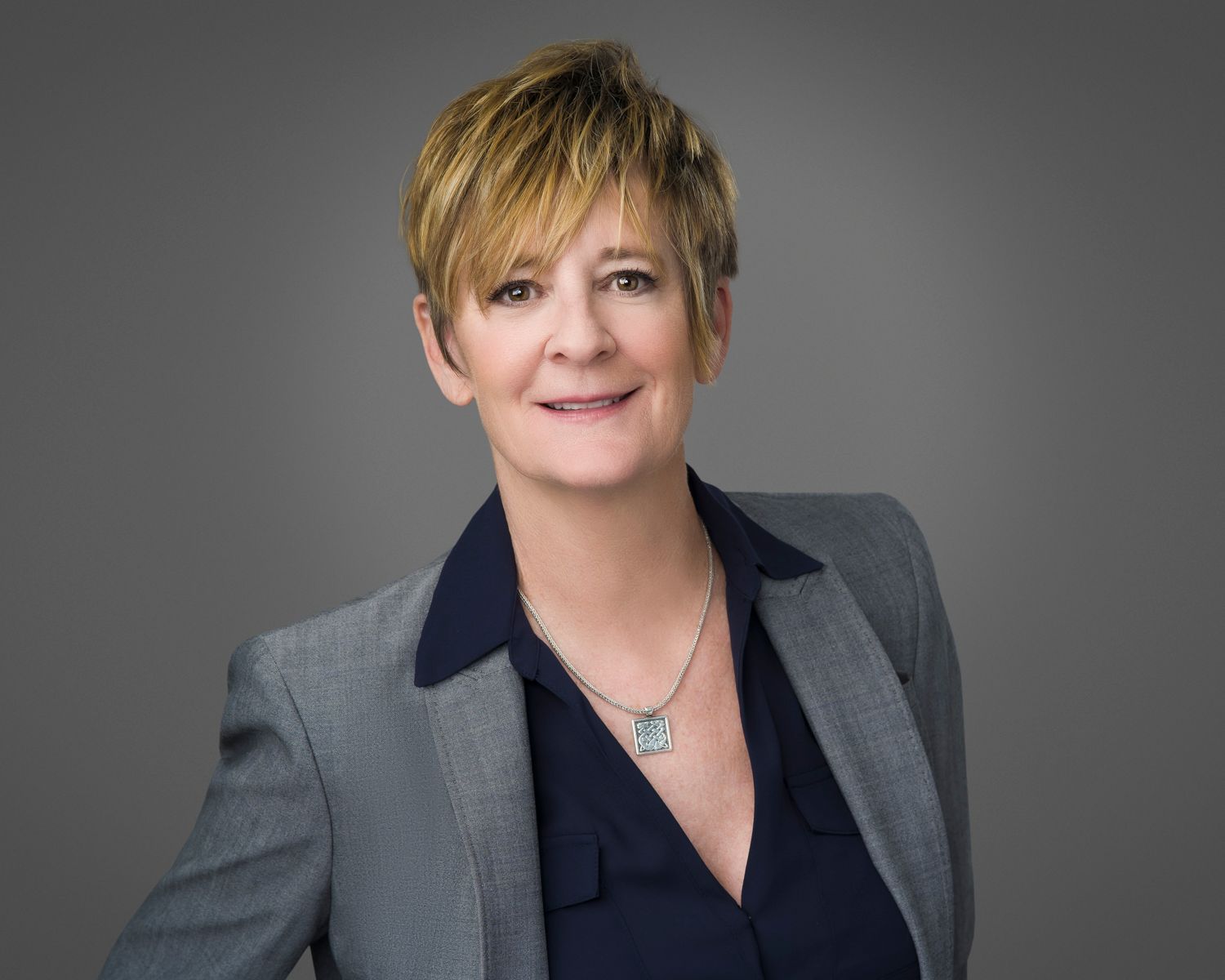 Professional portrait of a person with short blonde hair wearing grey blazer and navy top.