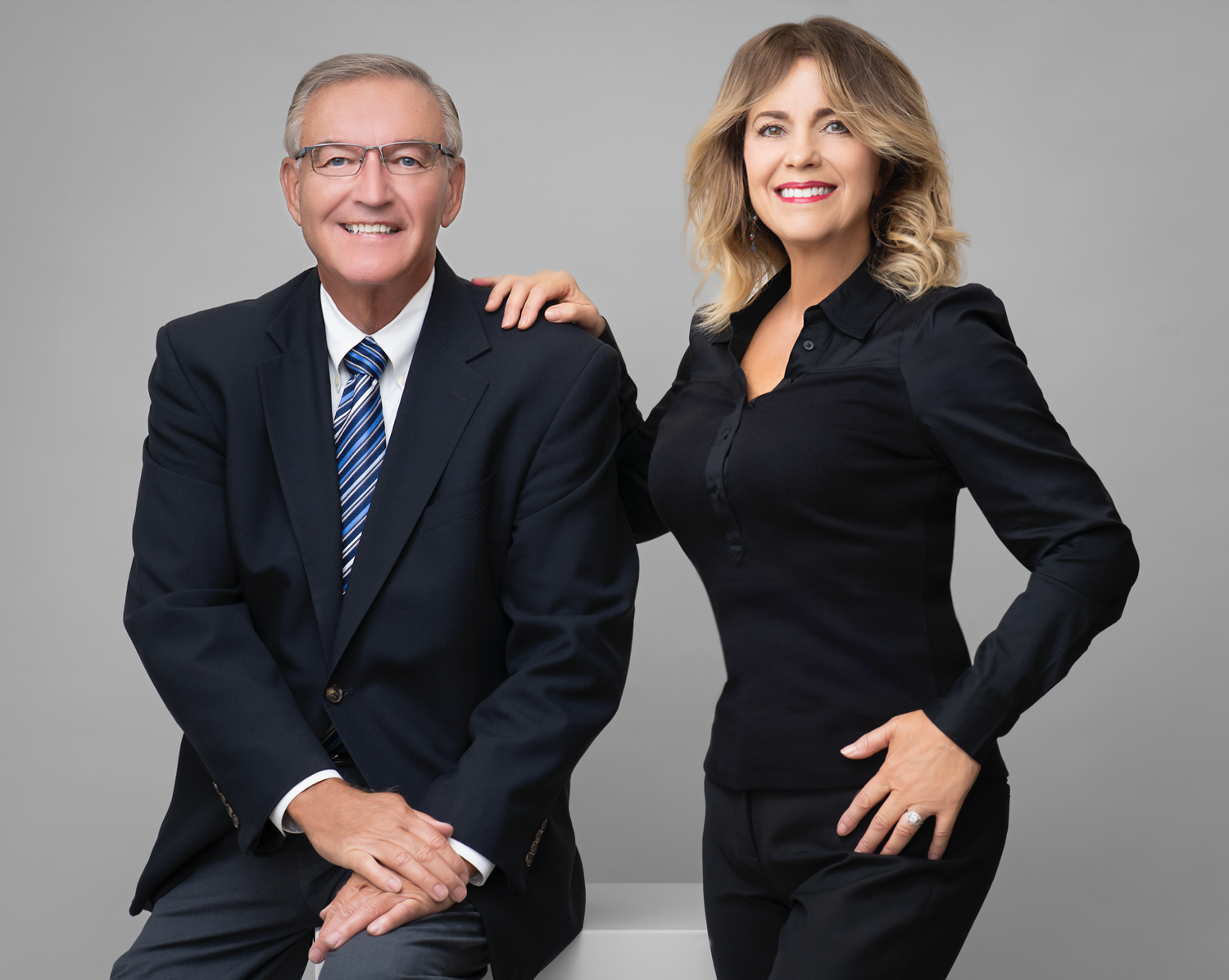 Professional business portrait of two colleagues in black attire against a gray studio background.