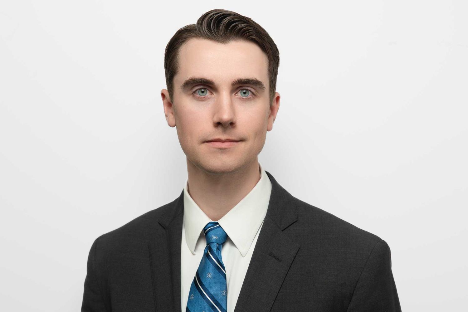 Young professional man in dark suit and blue striped tie poses for formal headshot against white background.
