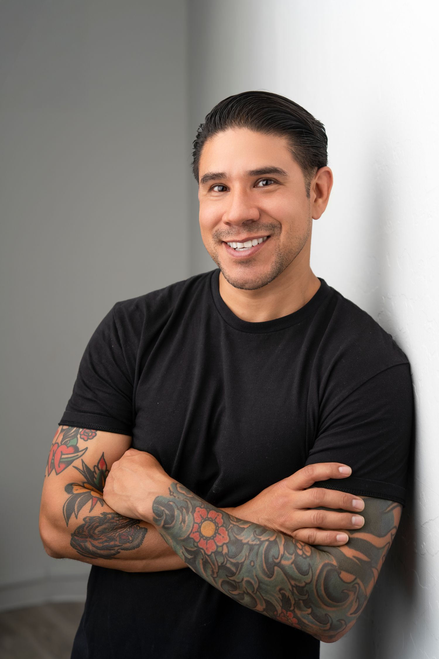 Professional portrait of a person in a black t-shirt with colorful sleeve tattoos smiling against a gray background.