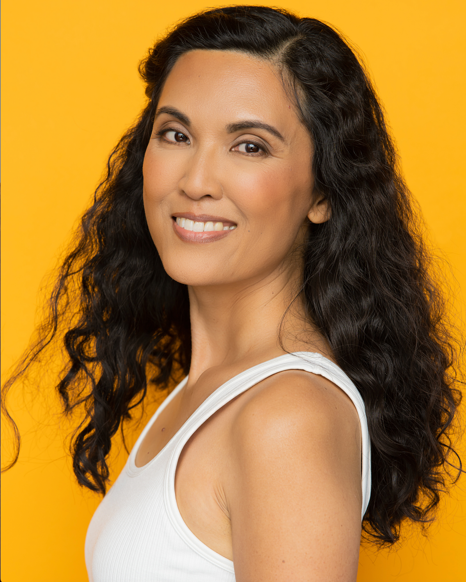 Woman with curly dark hair smiling against vibrant orange background wearing white sleeveless top.