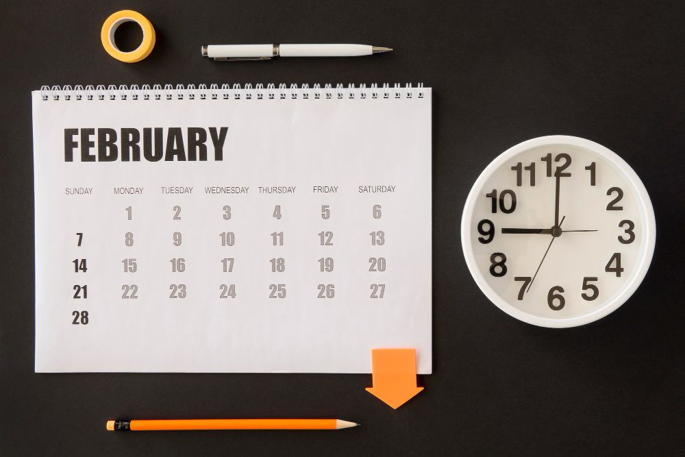 Desktop calendar showing February with clock and office supplies on dark background.