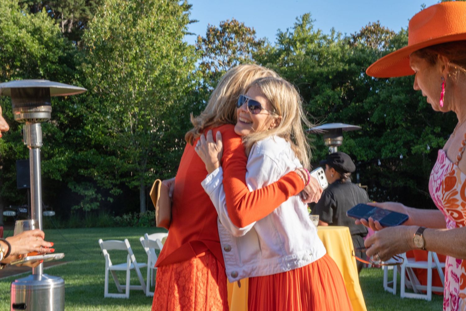 Two women hugging warmly at an outdoor garden party, dressed in vibrant orange outfits on a sunny day.
