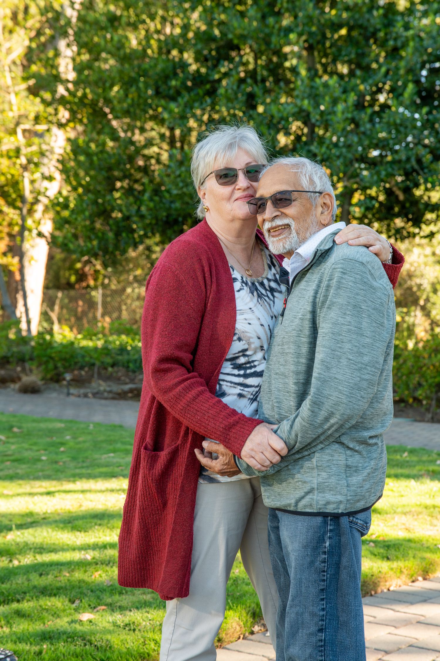 Senior couple embracing outdoors in a sunny garden, woman in red cardigan kissing mans cheek.