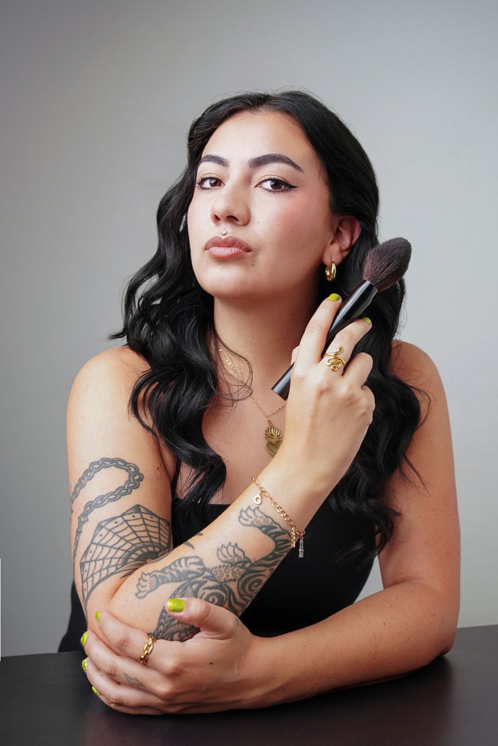 Professional beauty portrait with intricate arm tattoos, gold jewelry, and makeup brushes against neutral background.
