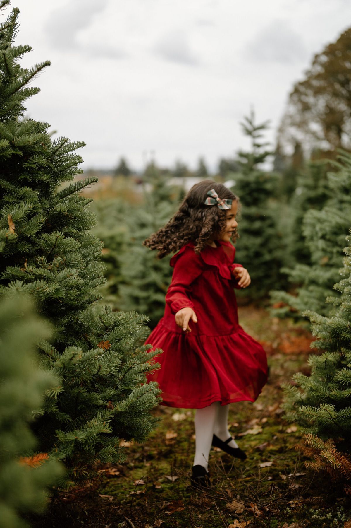 A young girl in a red dress twirls joyfully among rows of green Christmas trees at a tree farm.