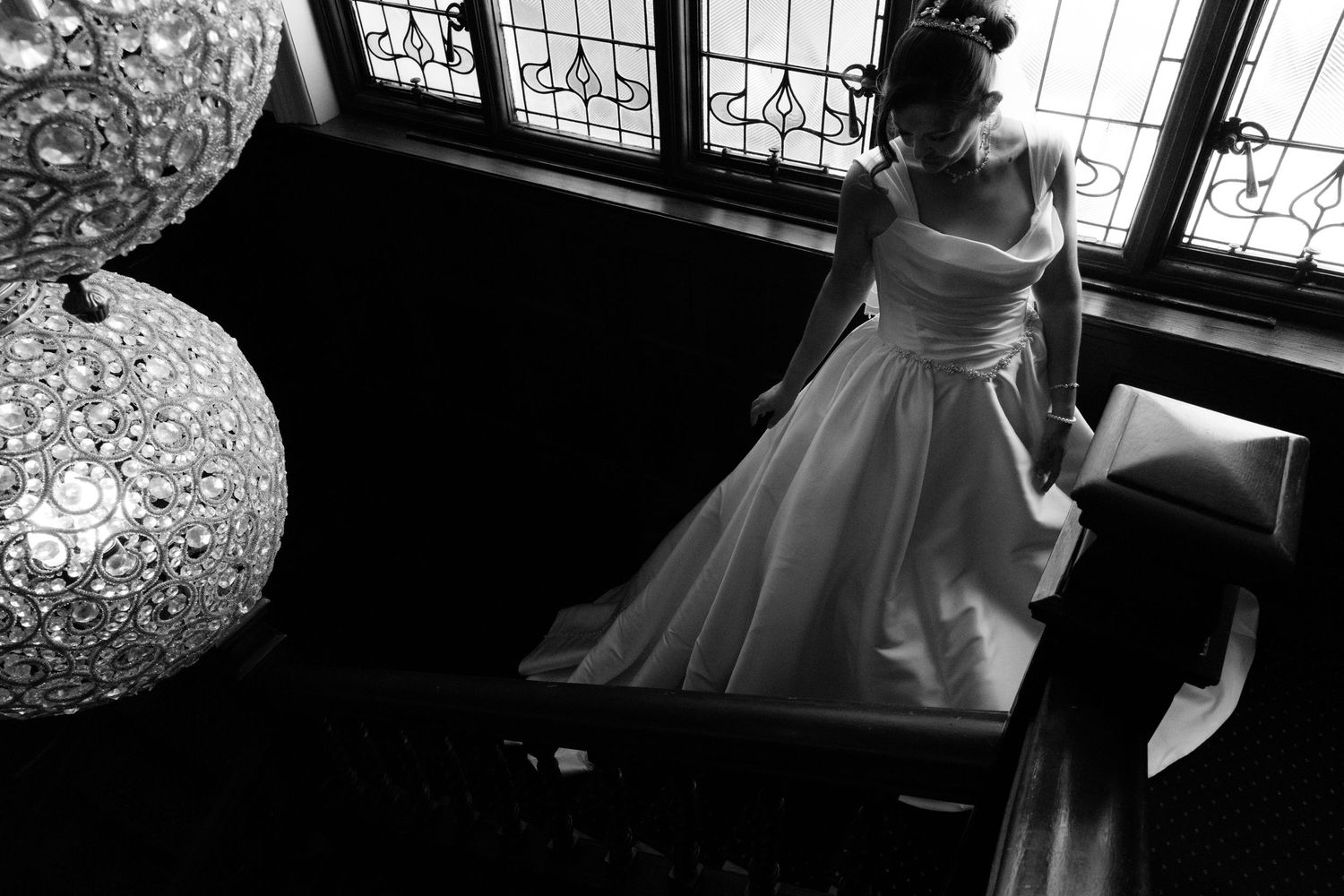 Black and white photo of a bride in a white ball gown standing near ornate windows and a decorative lamp.