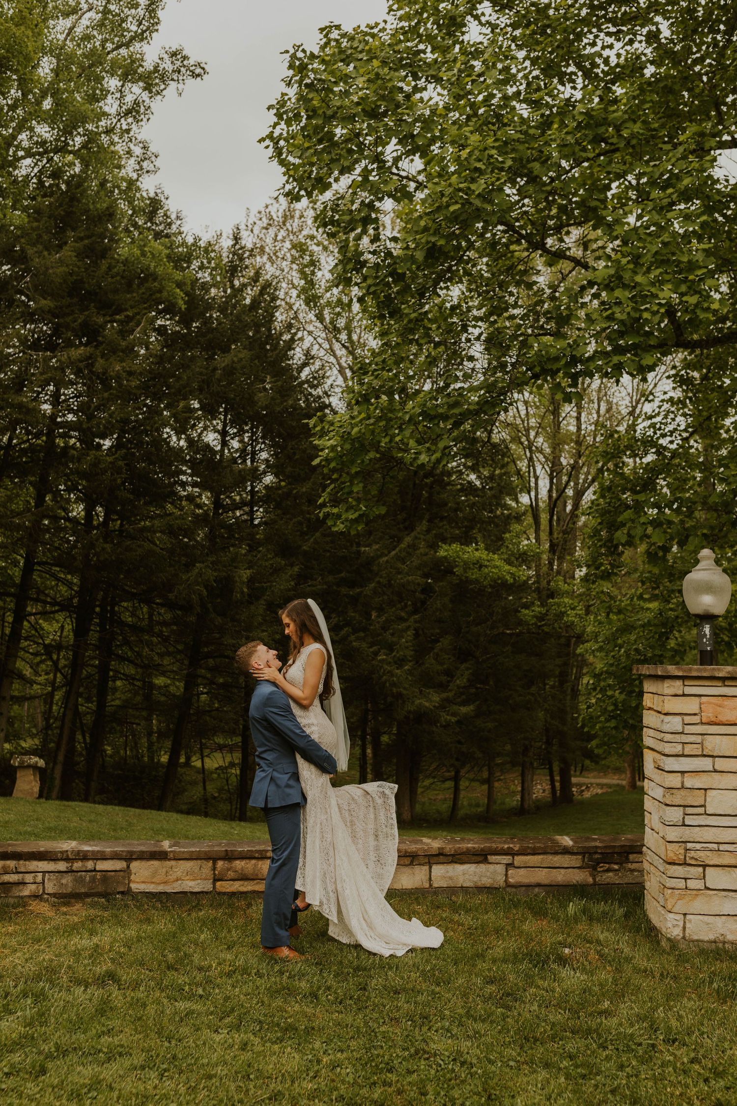 A romantic wedding moment captured outdoors with towering trees and a brick pillar in the background.