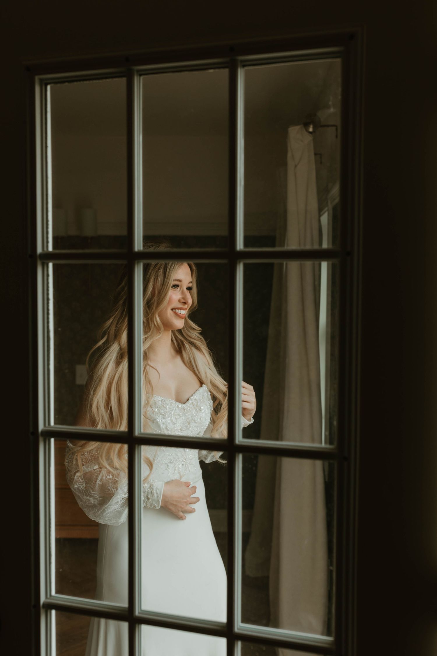 A bridal portrait through a divided window pane showing a woman in a lace wedding gown.