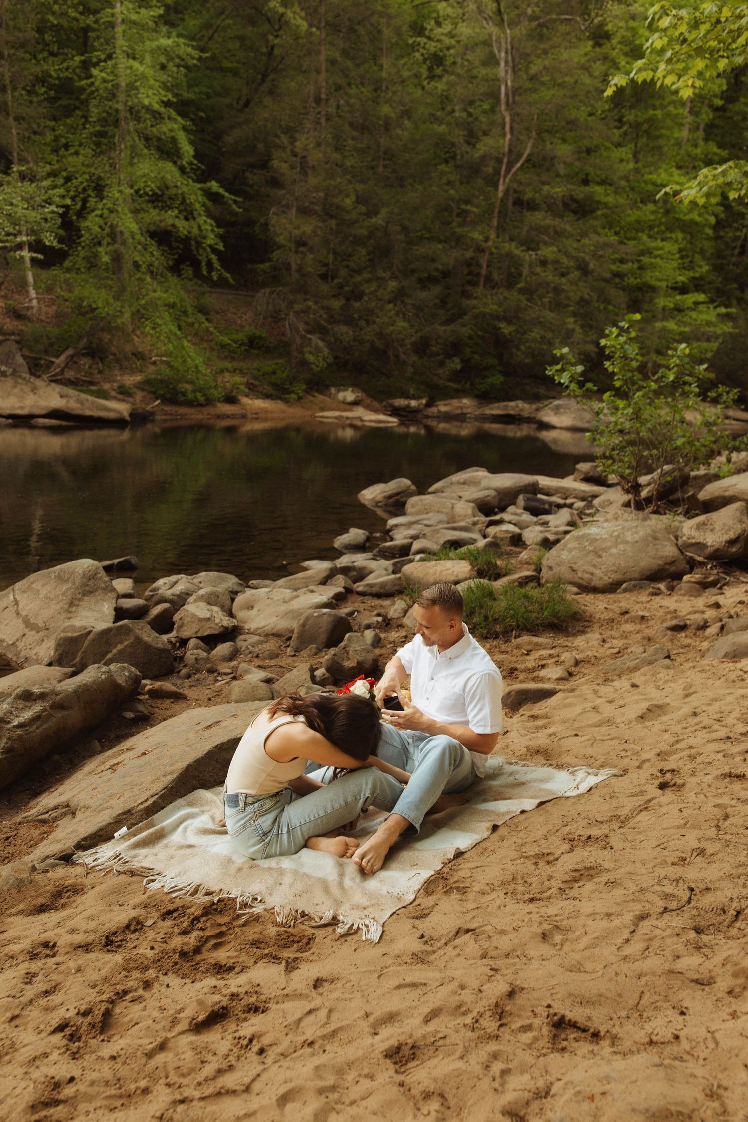 Two people share a peaceful moment on a blanket by a rocky riverbank.