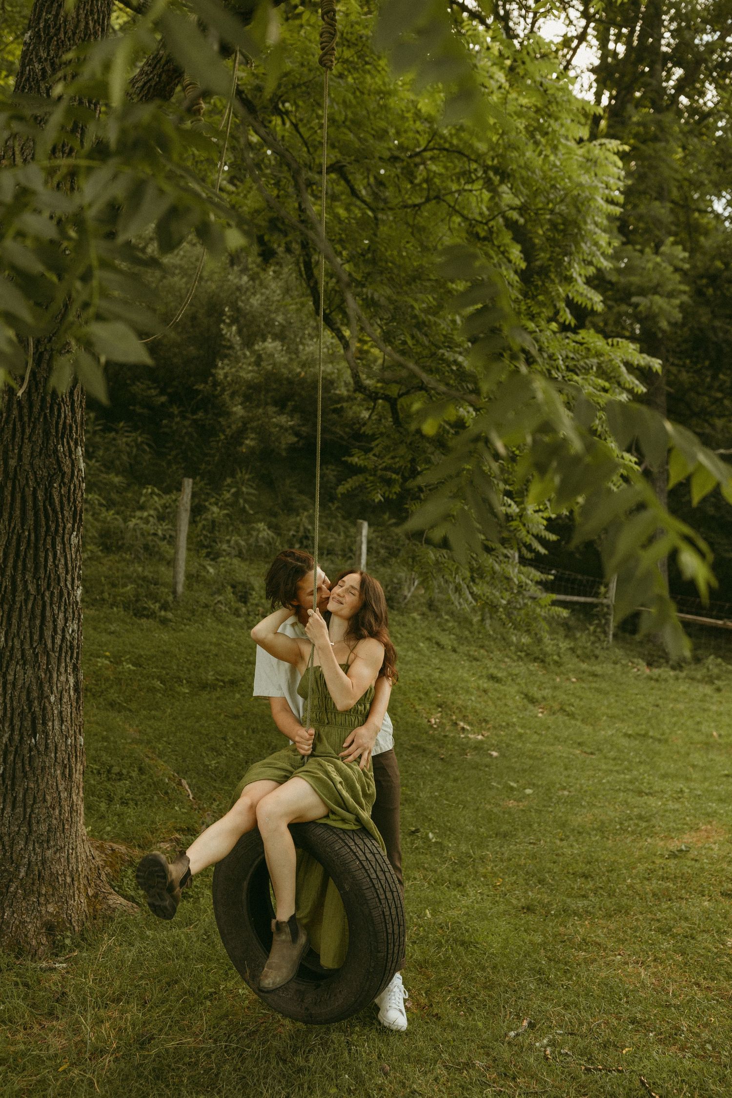 A couple enjoys a playful moment on a tire swing under green trees in a park.