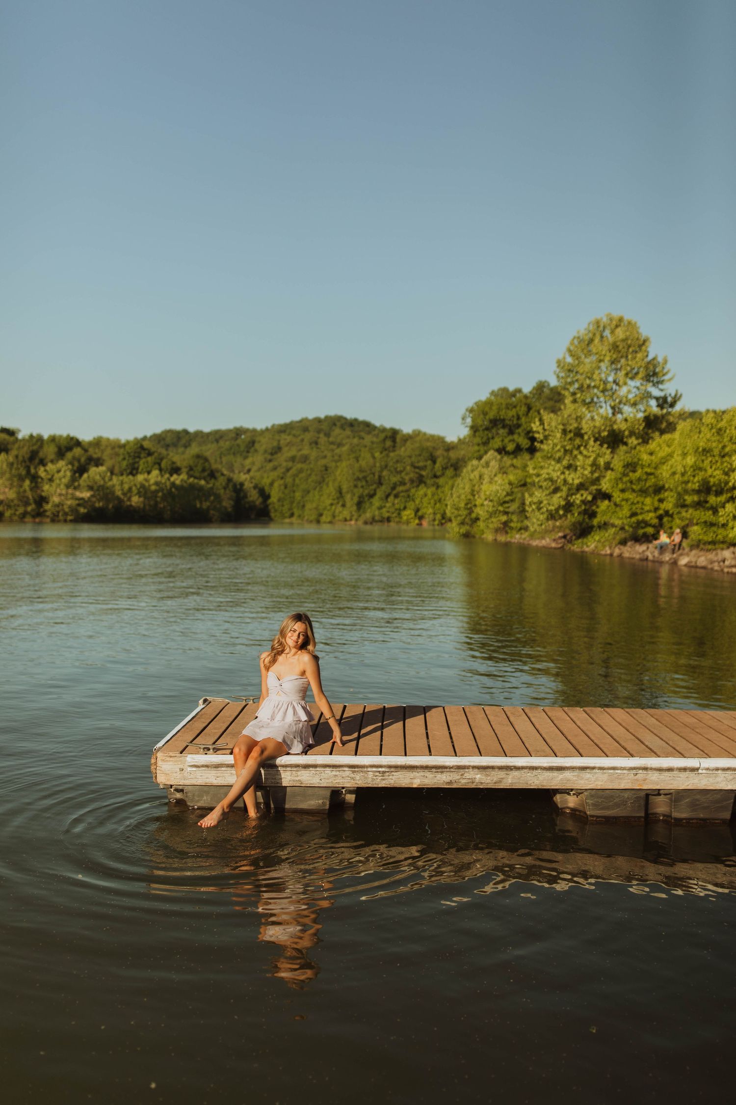 Someone in white sits on the edge of a wooden dock overlooking a tranquil lake.