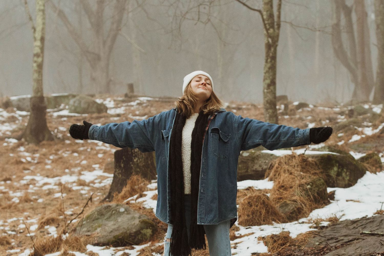 Person in denim jacket with arms outstretched in foggy winter forest.
