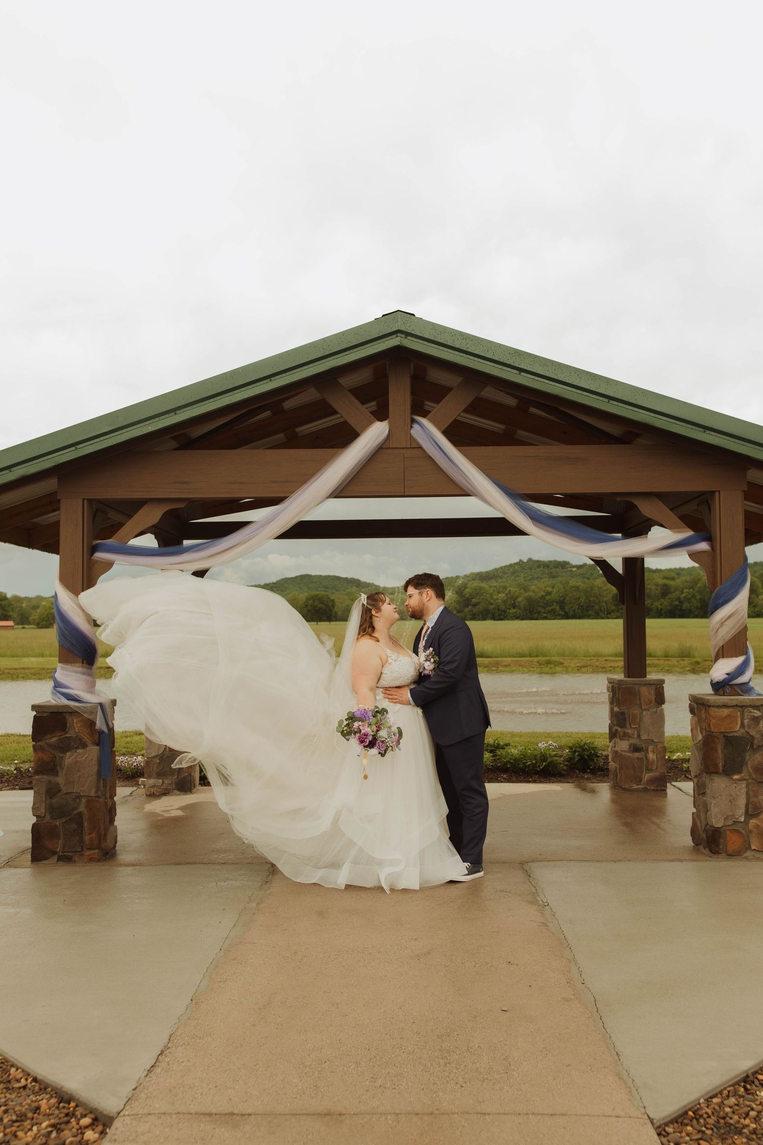 Wedding couple poses under wooden pavilion as bride's dress flows dramatically in the wind.