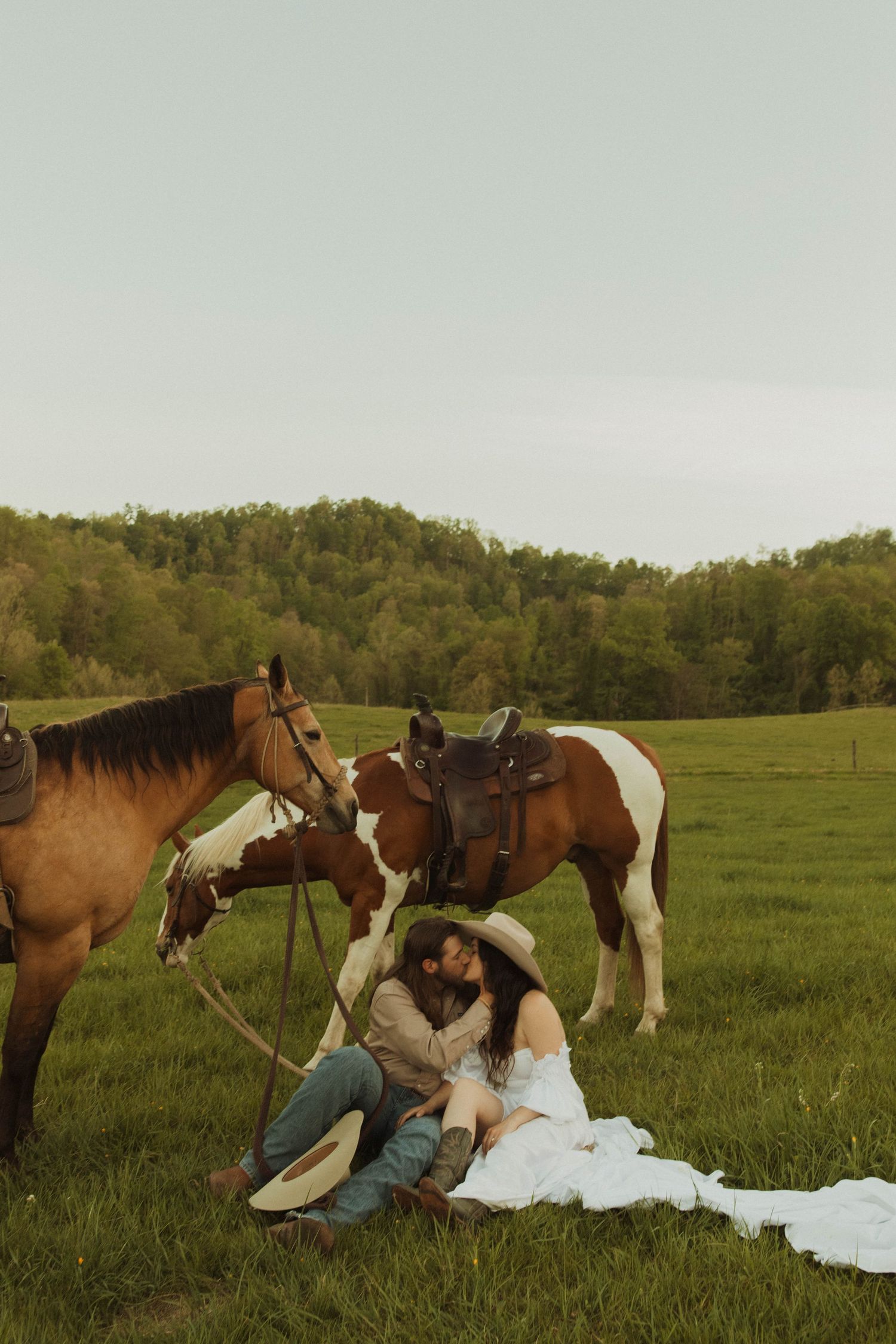 A couple rests on a blanket in a field with their horses nearby at sunset.
