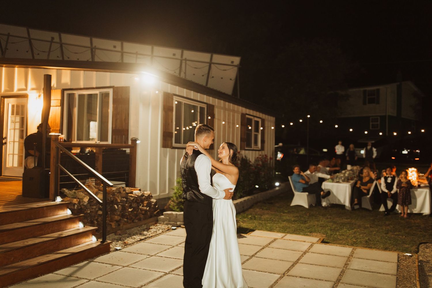 A couple shares their first dance on a patio under string lights at an evening wedding reception.