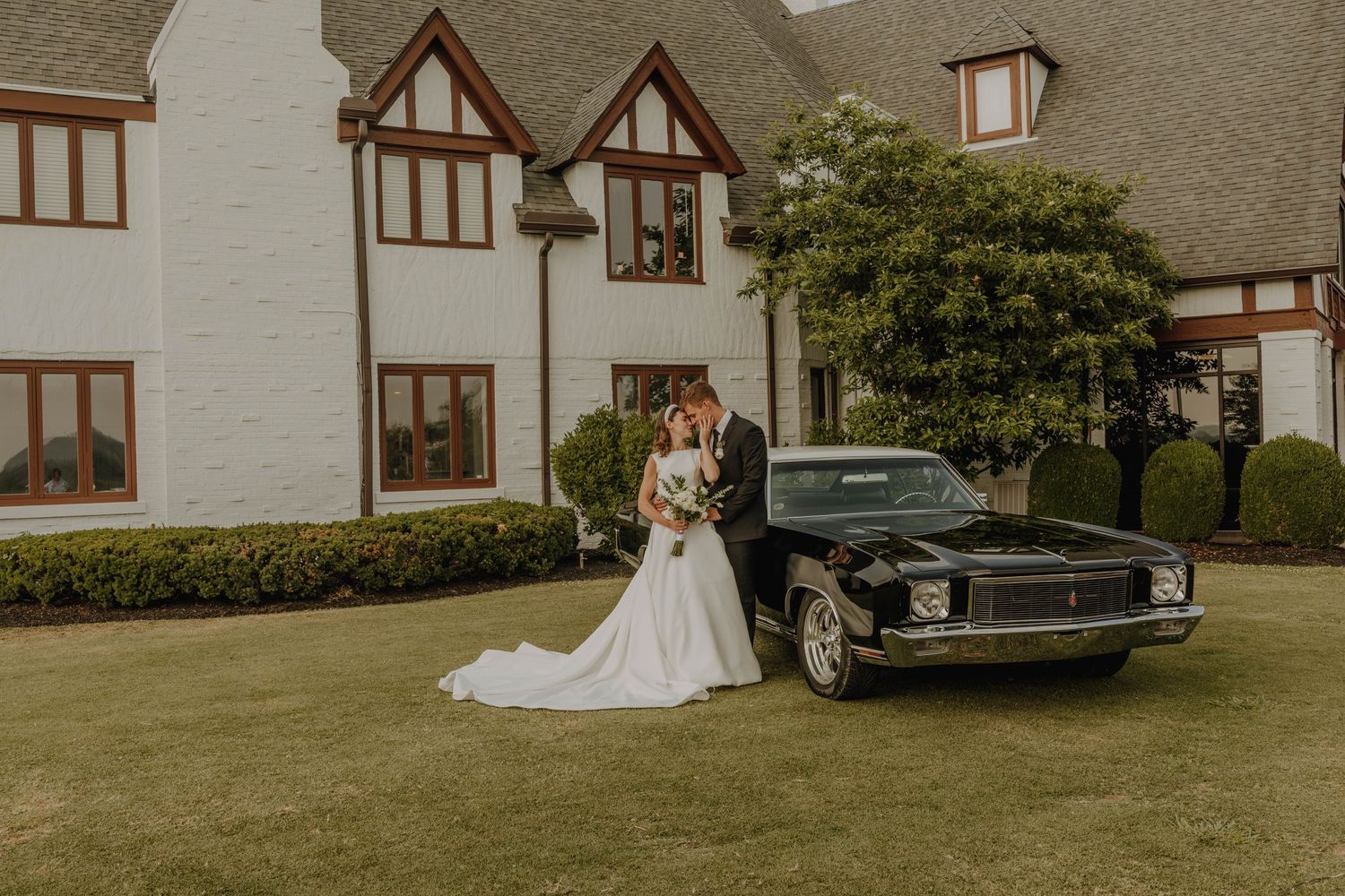 A wedding couple poses beside a vintage black car in front of a Tudor-style mansion.