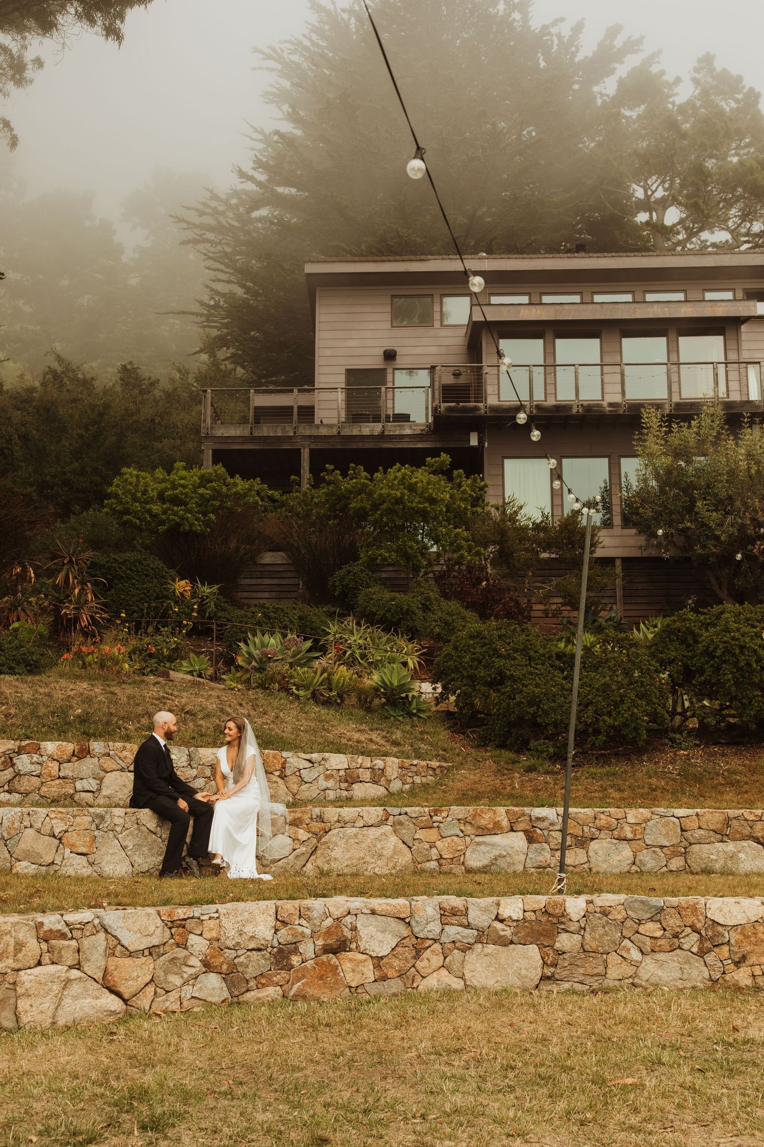 Couple sits on stone wall outside modern hillside home on misty afternoon.