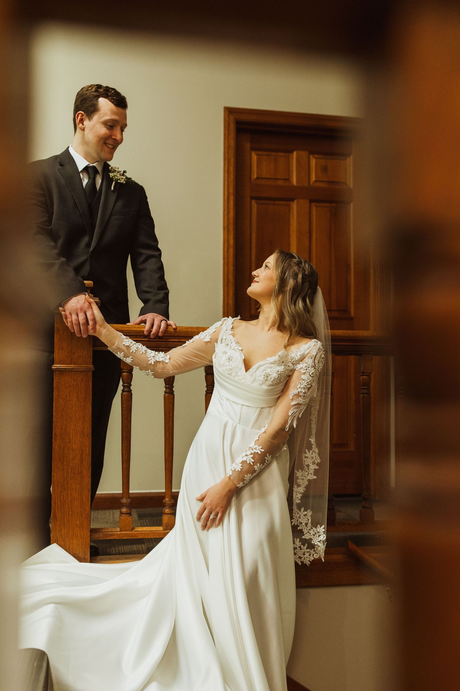 Elegant bride in lace gown poses on wooden staircase with groom in traditional suit.