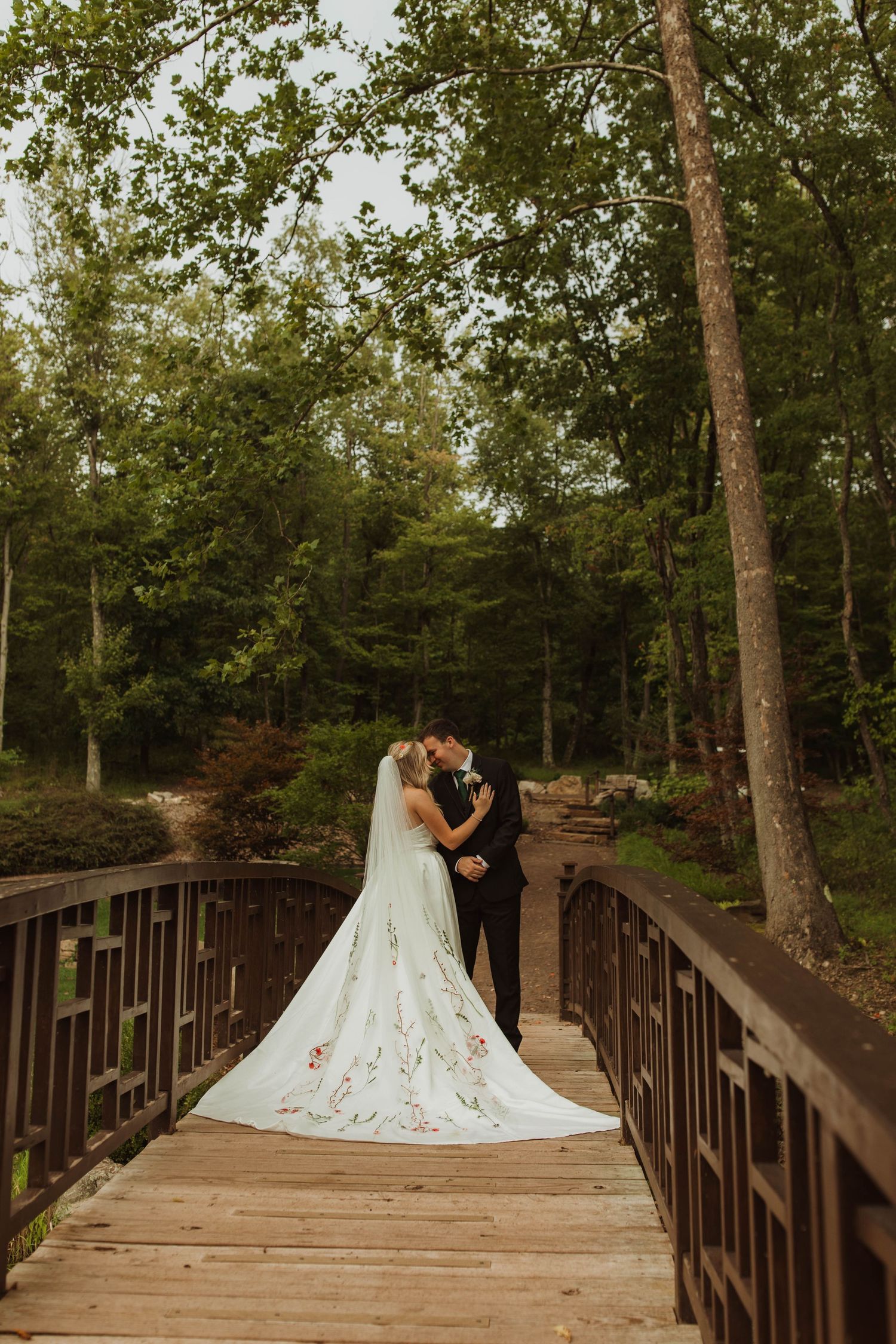 Wedding couple embracing on wooden bridge surrounded by lush forest scenery.