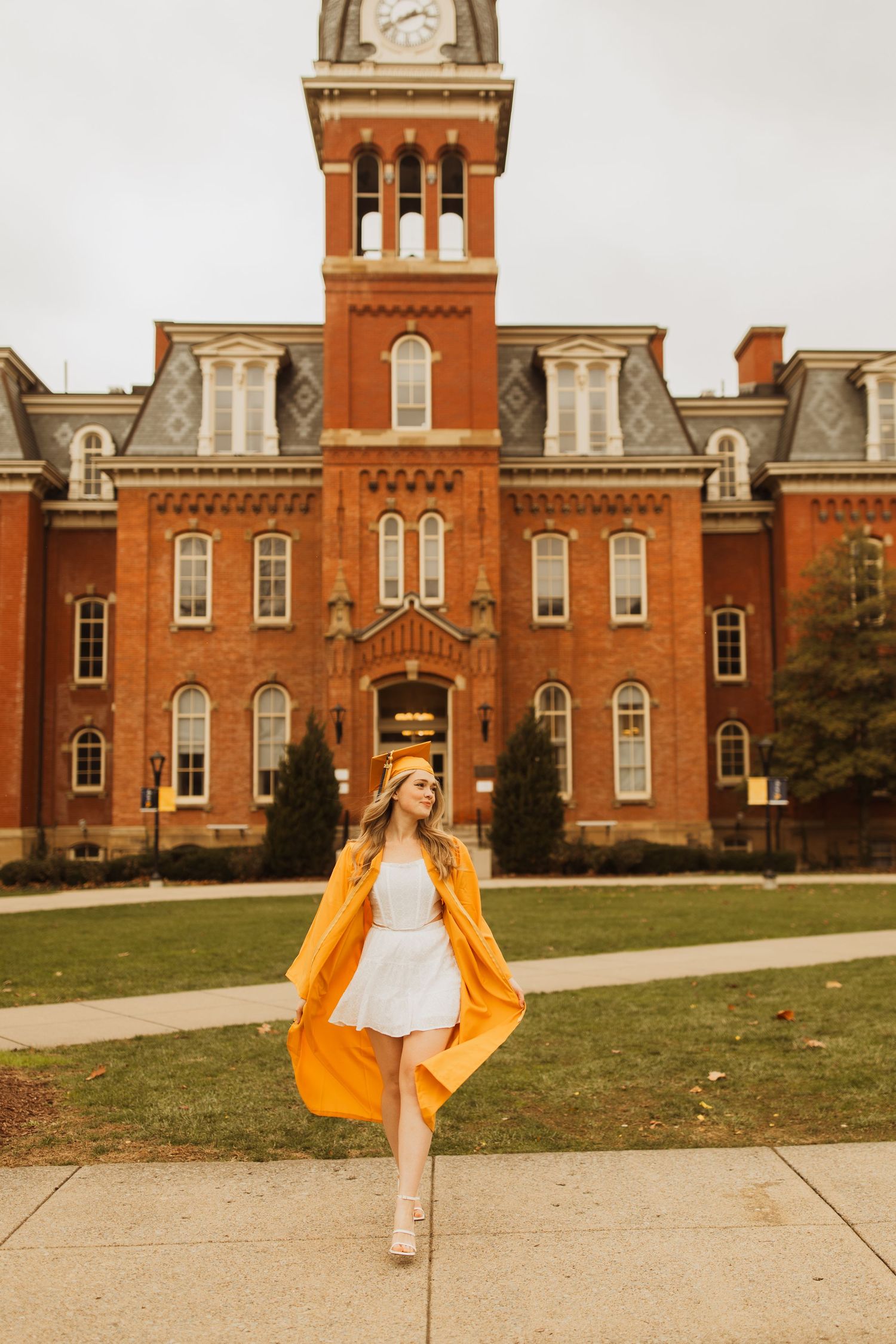 Person in orange cape and white dress posing in front of historic red brick university building.