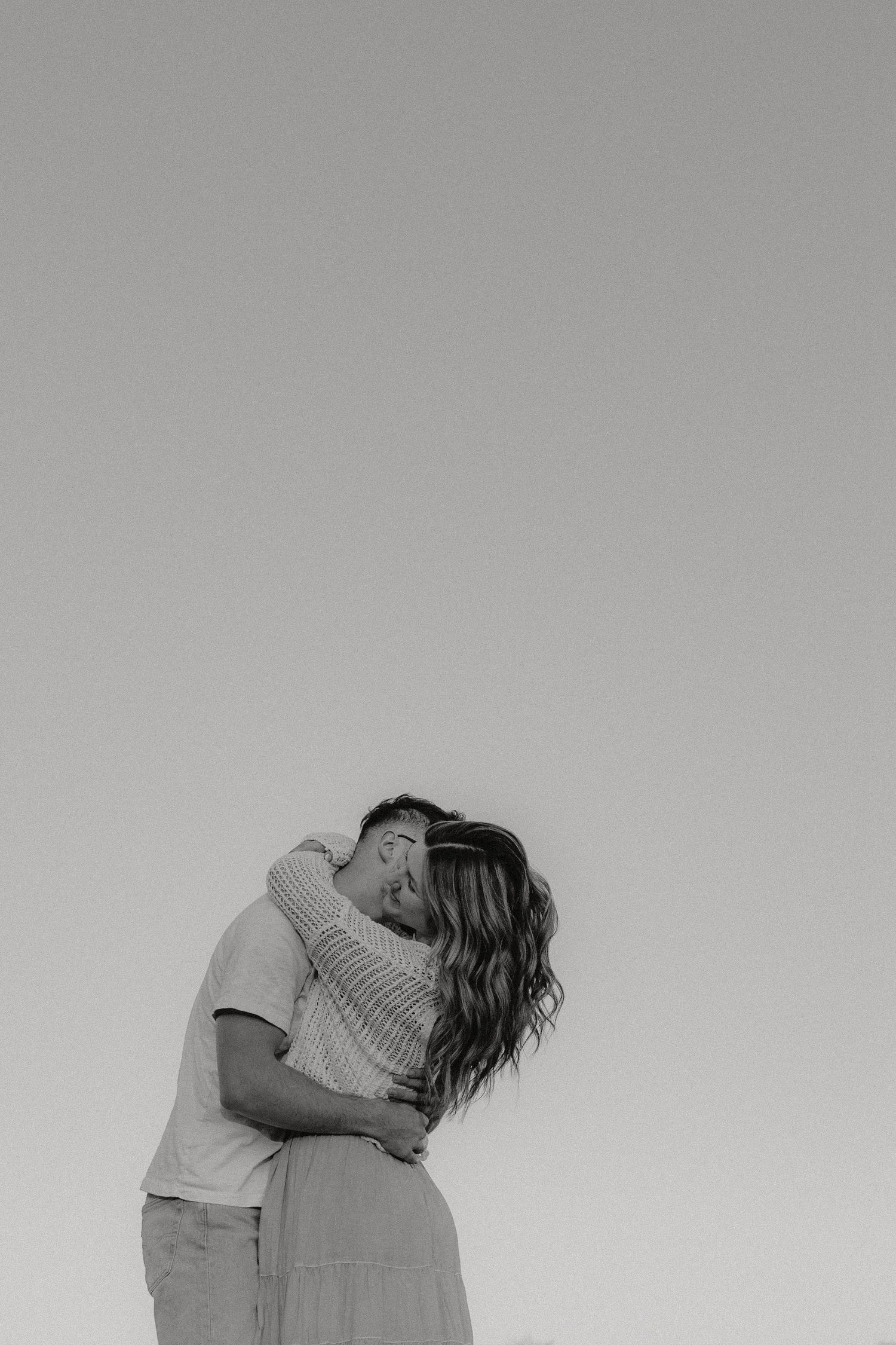 A romantic black and white portrait of a couple embracing against a minimalist sky background.
