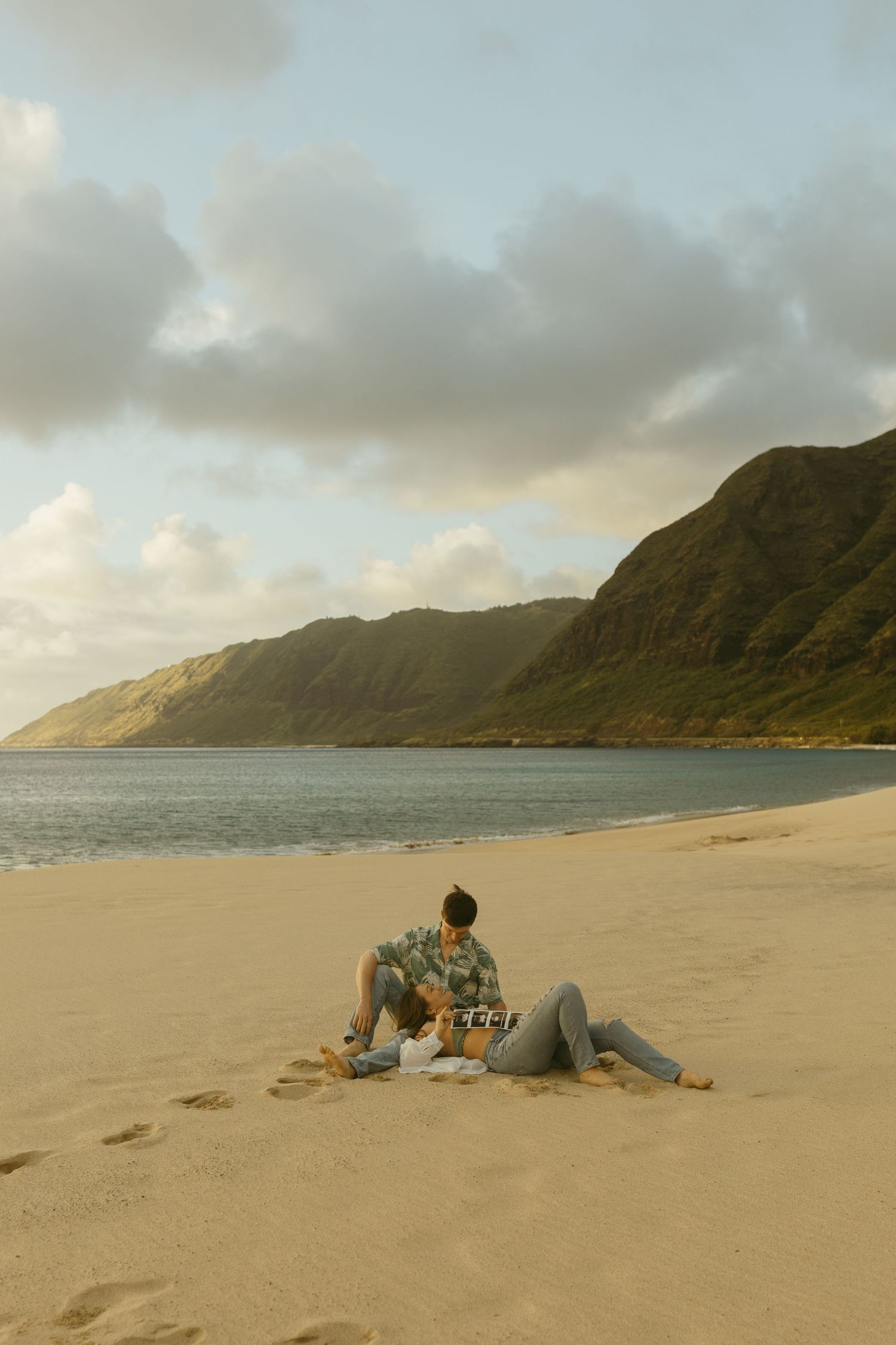 Two people relax on sandy beach with majestic coastal mountains in background.