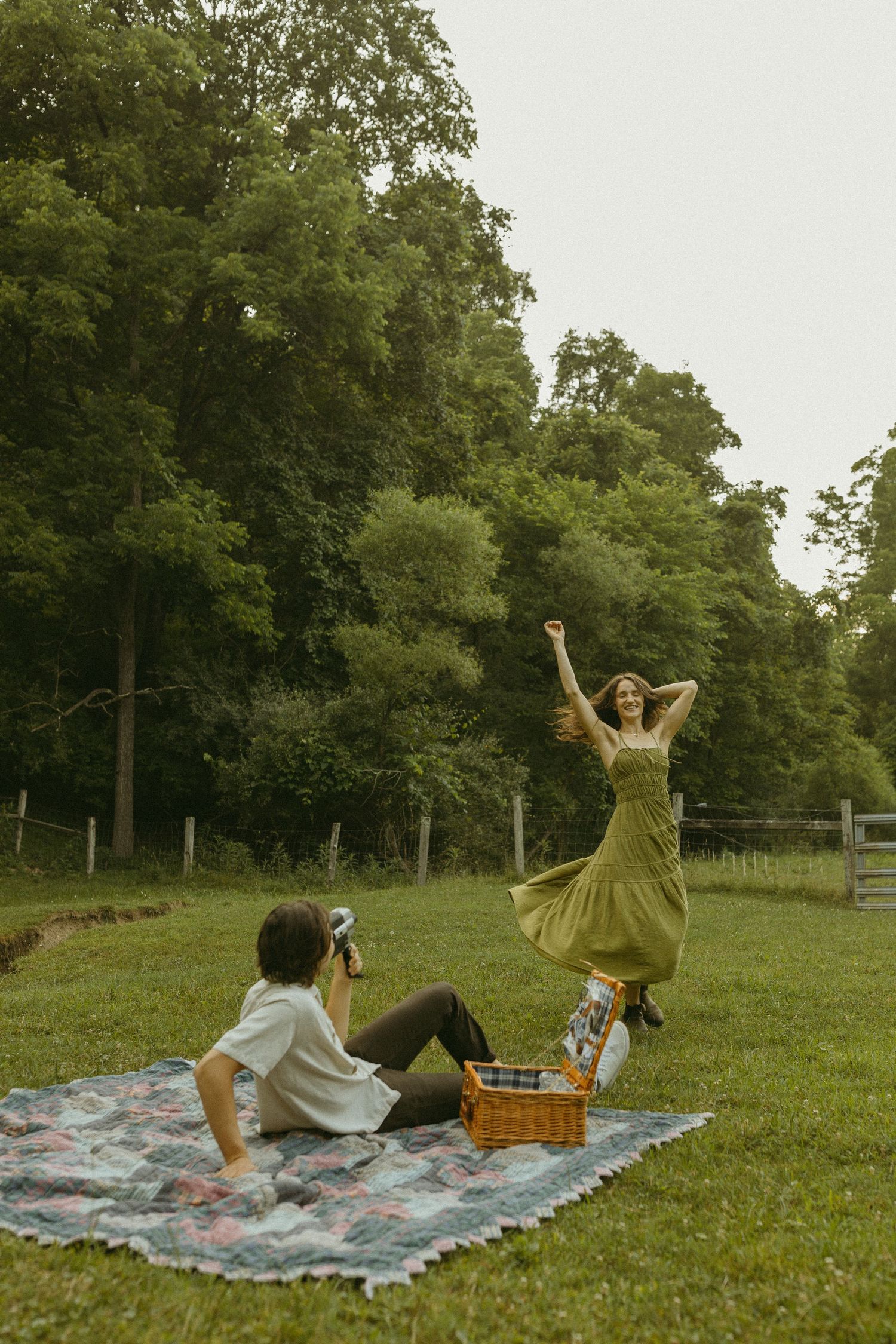 Person leaps joyfully in a flowing olive dress while another captures the moment during a picnic.