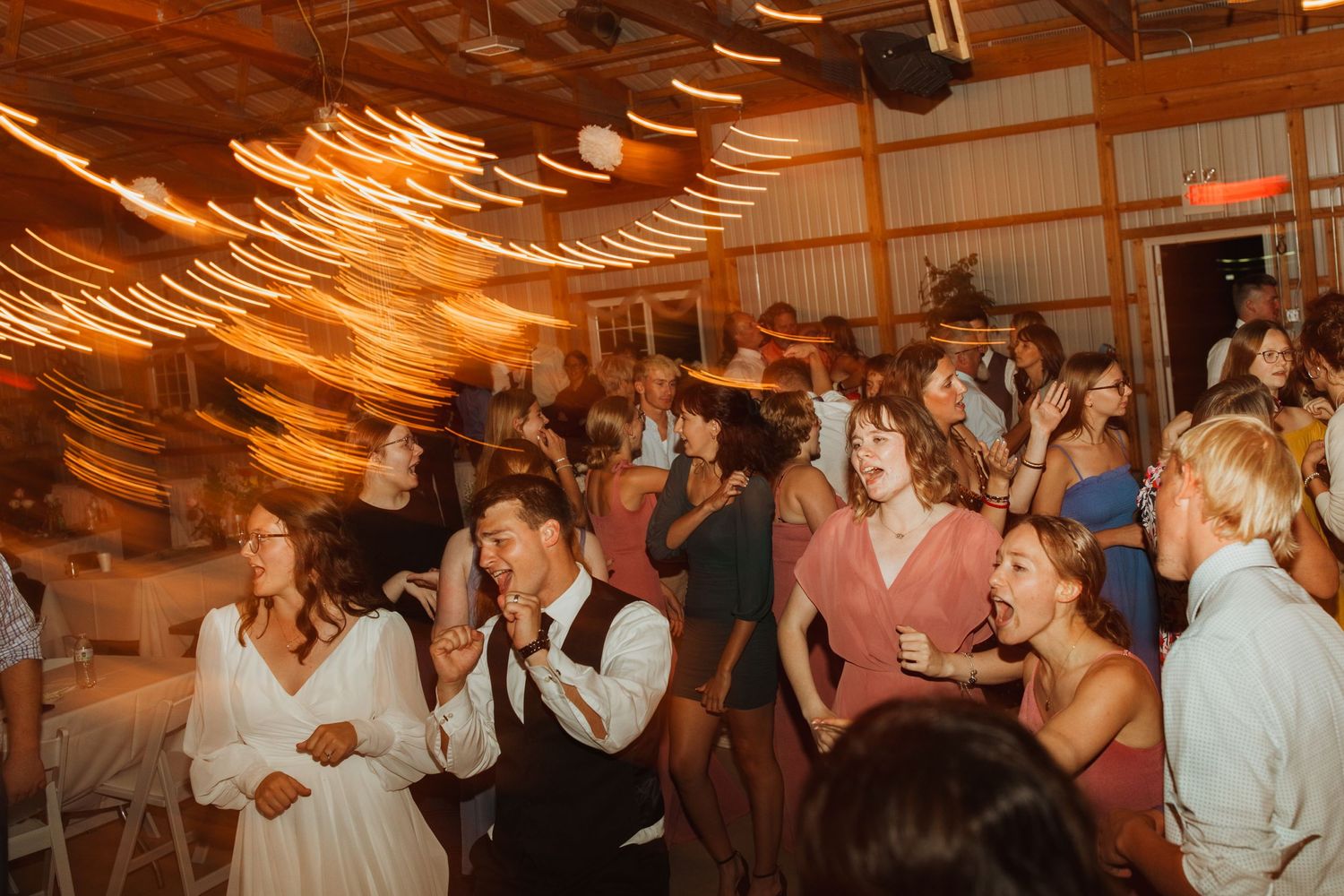 Wedding reception party with dancing guests under warm string lights in a barn.